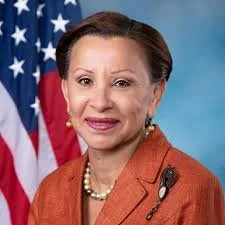 Portrait of Congresswomen Nydia Velazquez in an orange blazer, wearing a pearl necklace and earrings, with an American flag in the background.