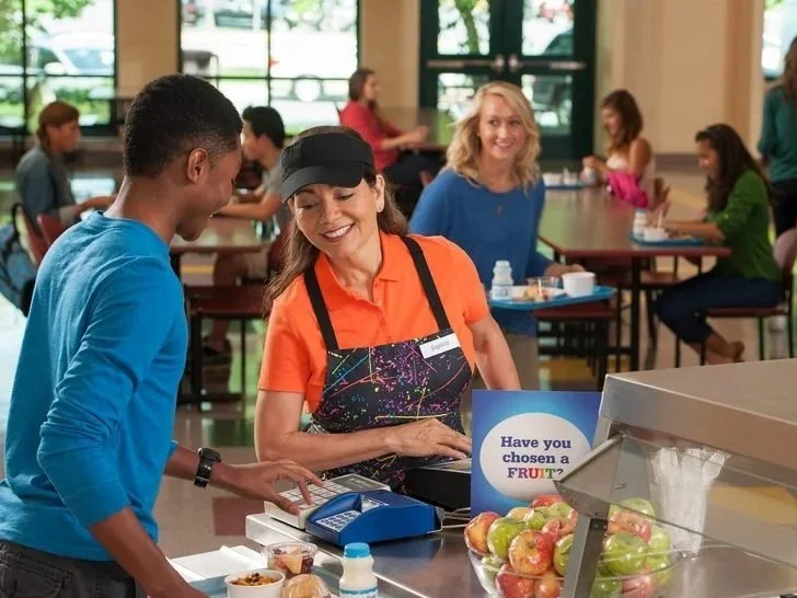 A woman in an orange shirt and apron is smiling and working at a fruit stand or cafeteria register, with a young man in a blue shirt and black cap paying. In the background, there are other people sitting at tables, some eating and talking, inside a bright, modern space with large windows.