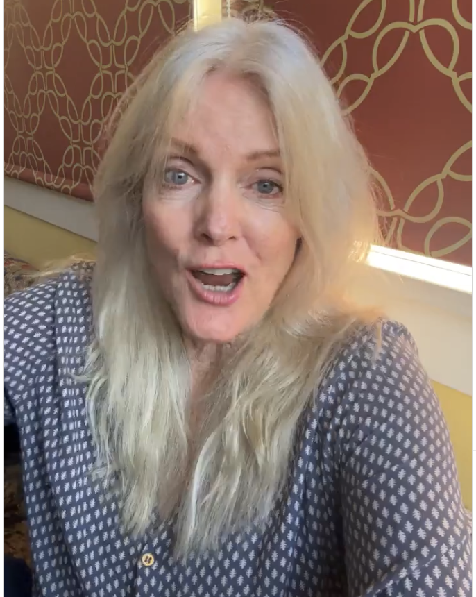 Maggie Baird, mother of Billie Eilish, wearing a grey patterned blouse, speaking or smiling in a brightly lit room with red and gold patterned decor on the wall behind her after interviewing the Plant Powered Youth Fellowship
