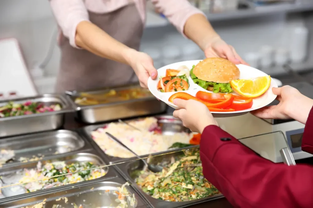 Person serving a burger with lettuce, tomato, and lemon wedge to a customer at a cafeteria or buffet-style food service counter.