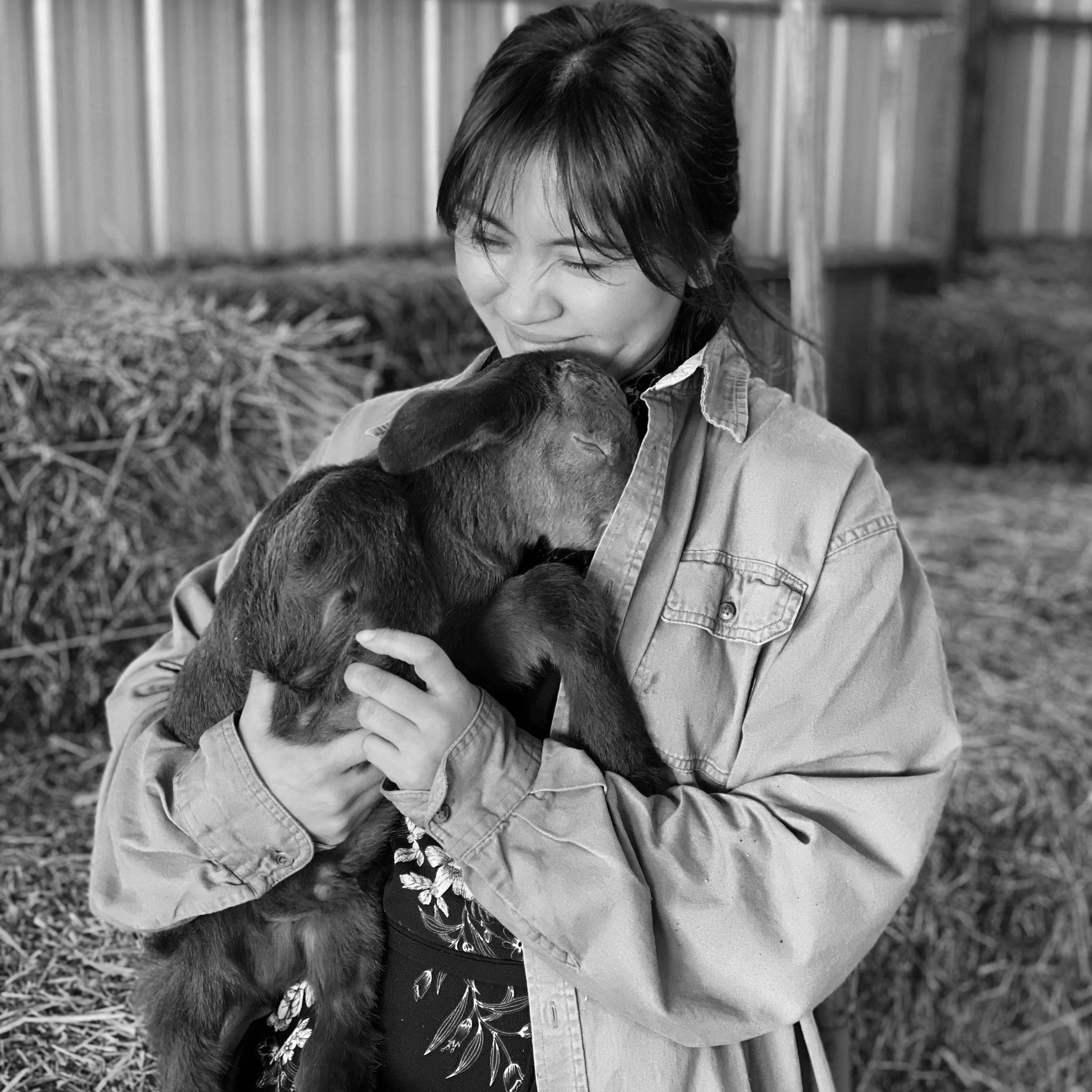 A woman holding a baby goat, gently pressing her face against its head in an outdoor farm setting.
