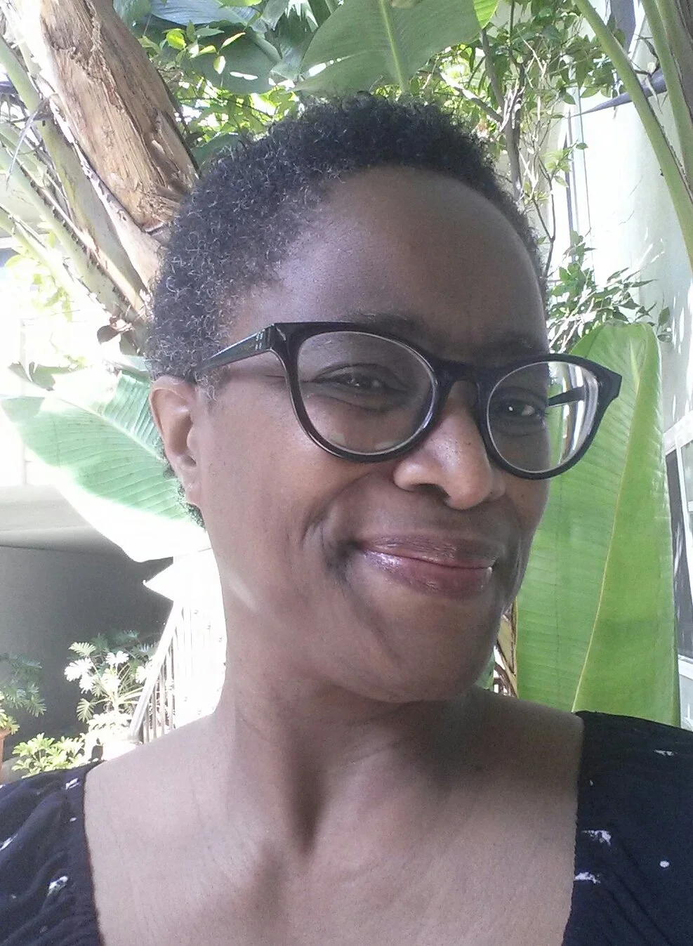 A woman with short curly hair wearing black glasses, smiling, with large green leaves and a tree in the background.