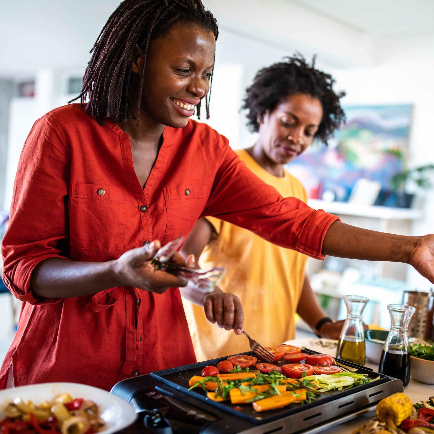 Two women cooking vegetables on a grill in a bright room.