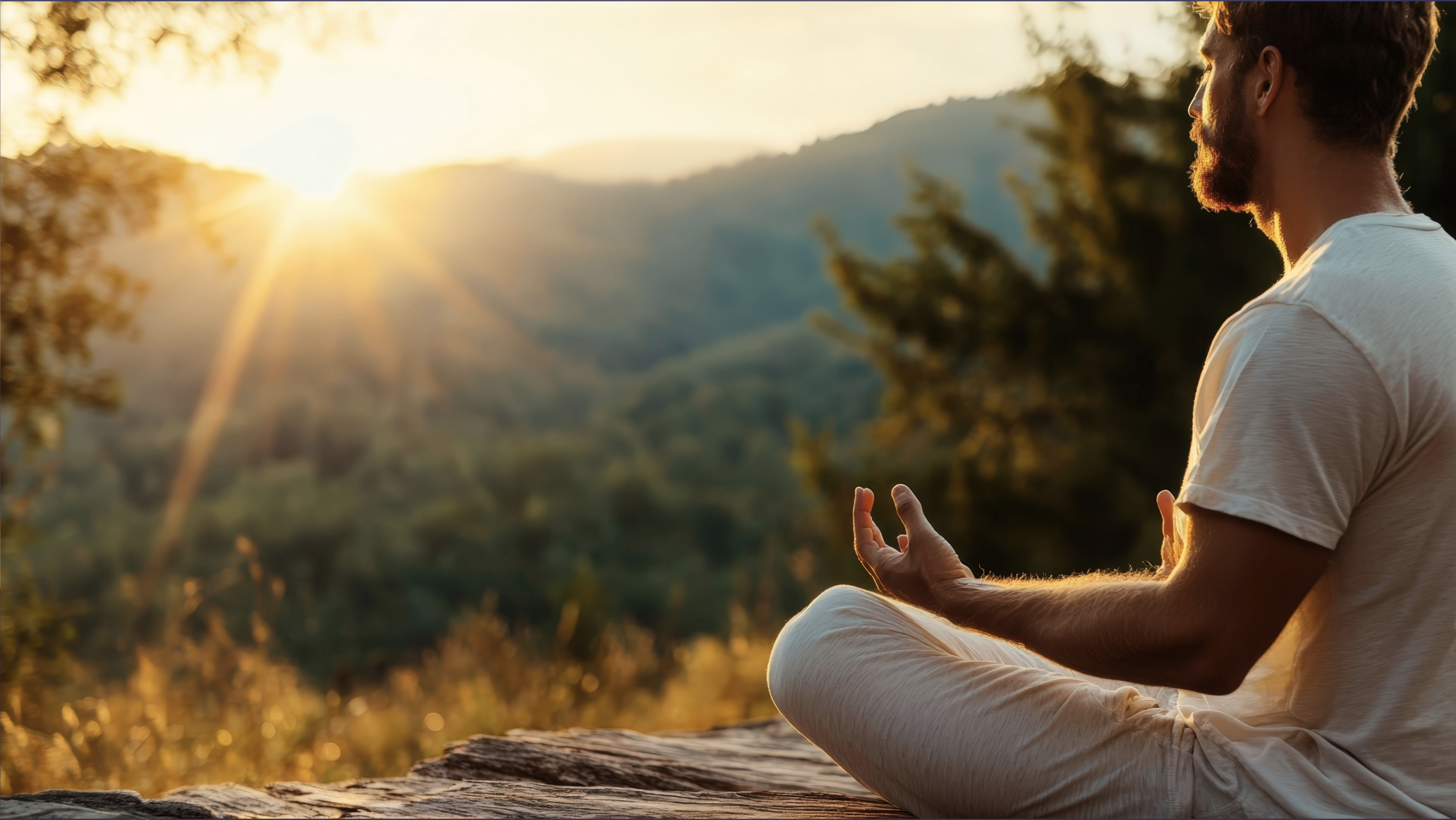 A man practicing yoga or meditation outdoors during sunset, sitting cross-legged on a wooden surface with a scenic mountain and forest background.