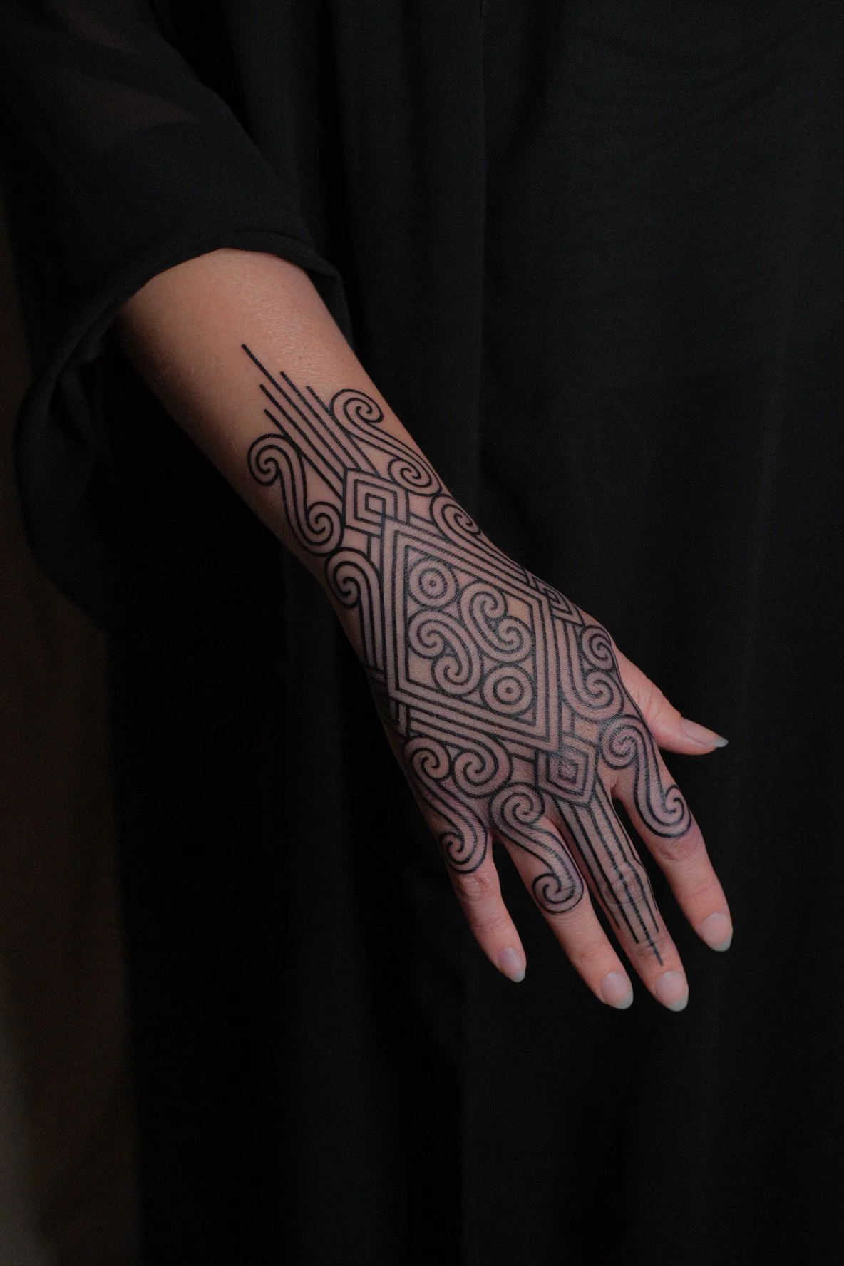 Tatouage noir complexe et géométrique sur le bras et la main d'une personne, avec un fond sombre.