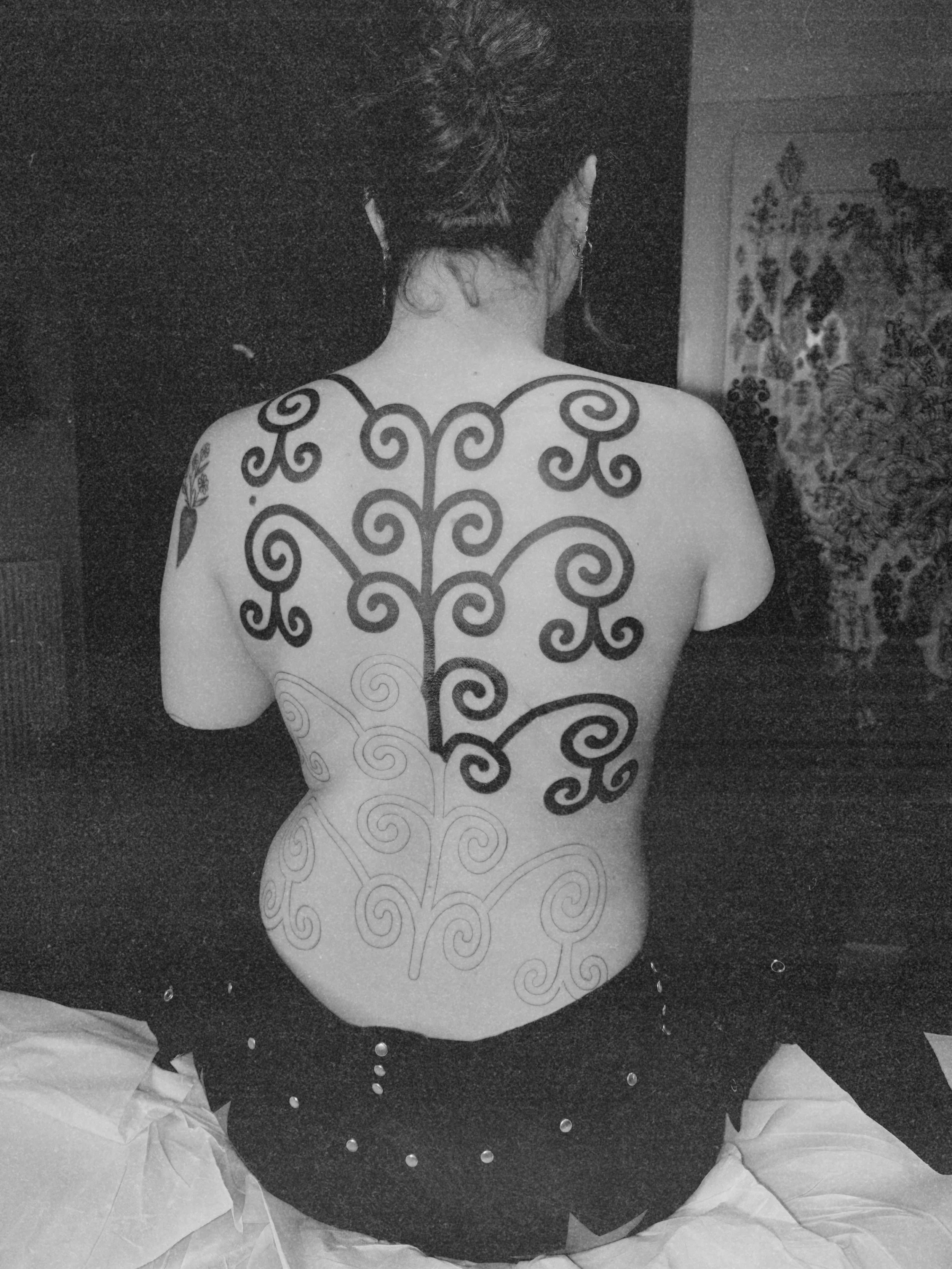 Une personne vue de dos avec un tatouage noir et un piercing à l'oreille, assise sur un lit, portant une jupe blanche avec des papillons géants et un haut noir, entourée de papier de soie blanc.