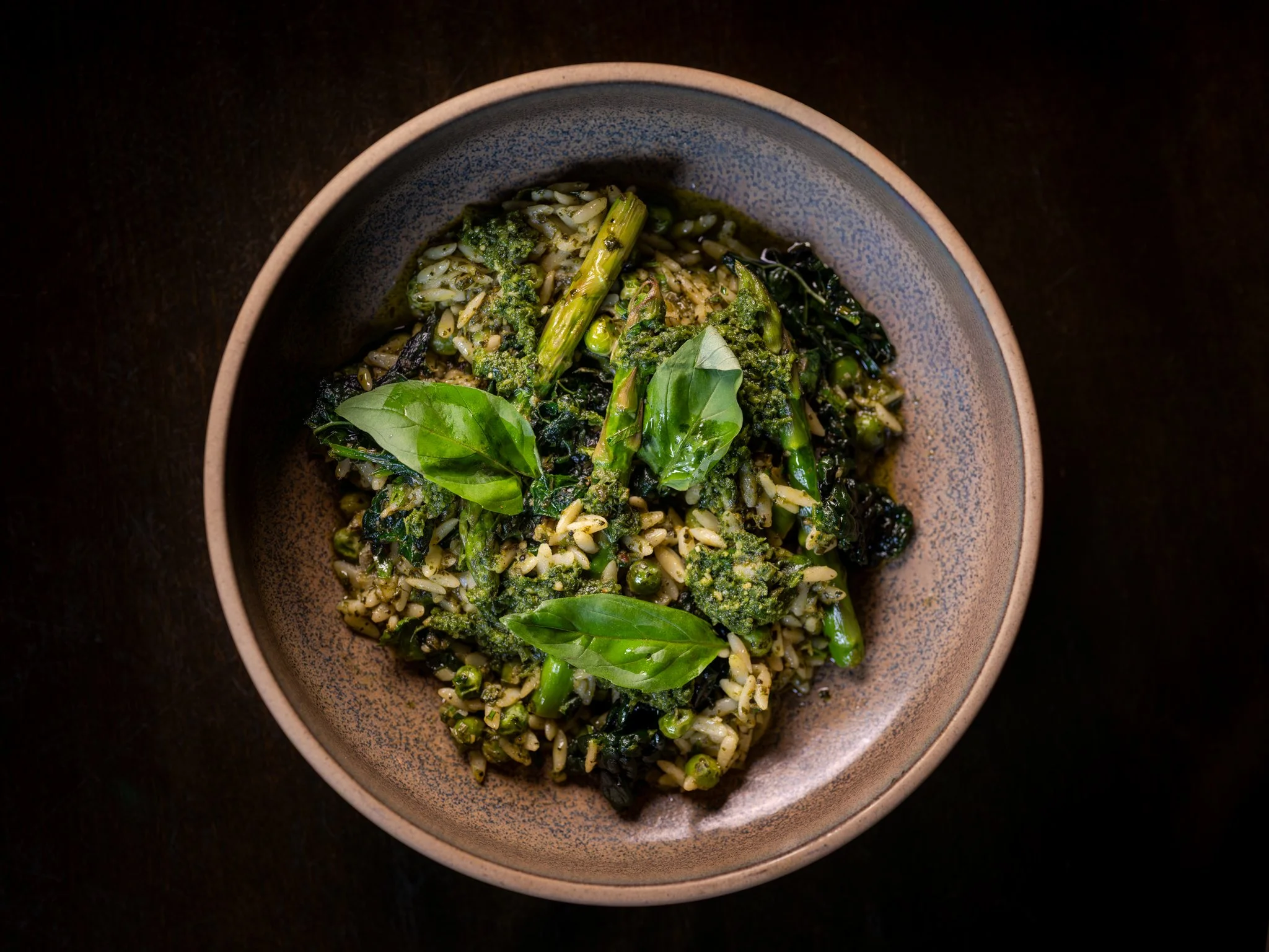 A bowl of cooked rice mixed with green vegetables, topped with fresh basil leaves, on a dark surface.
