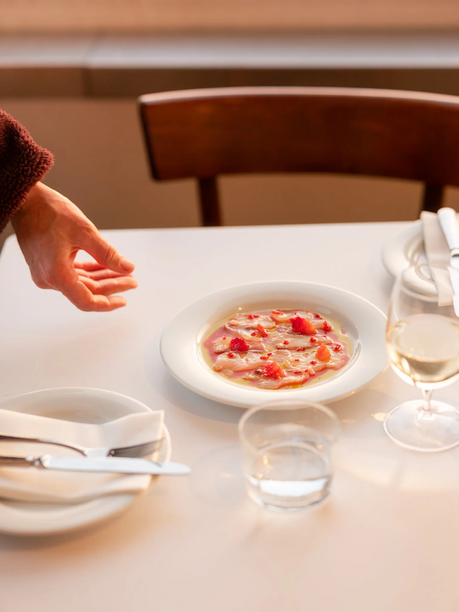 A table set with white plates, silverware, a glass of white wine, and a soup or appetizer with pink and white ingredients in a restaurant or dining setting.