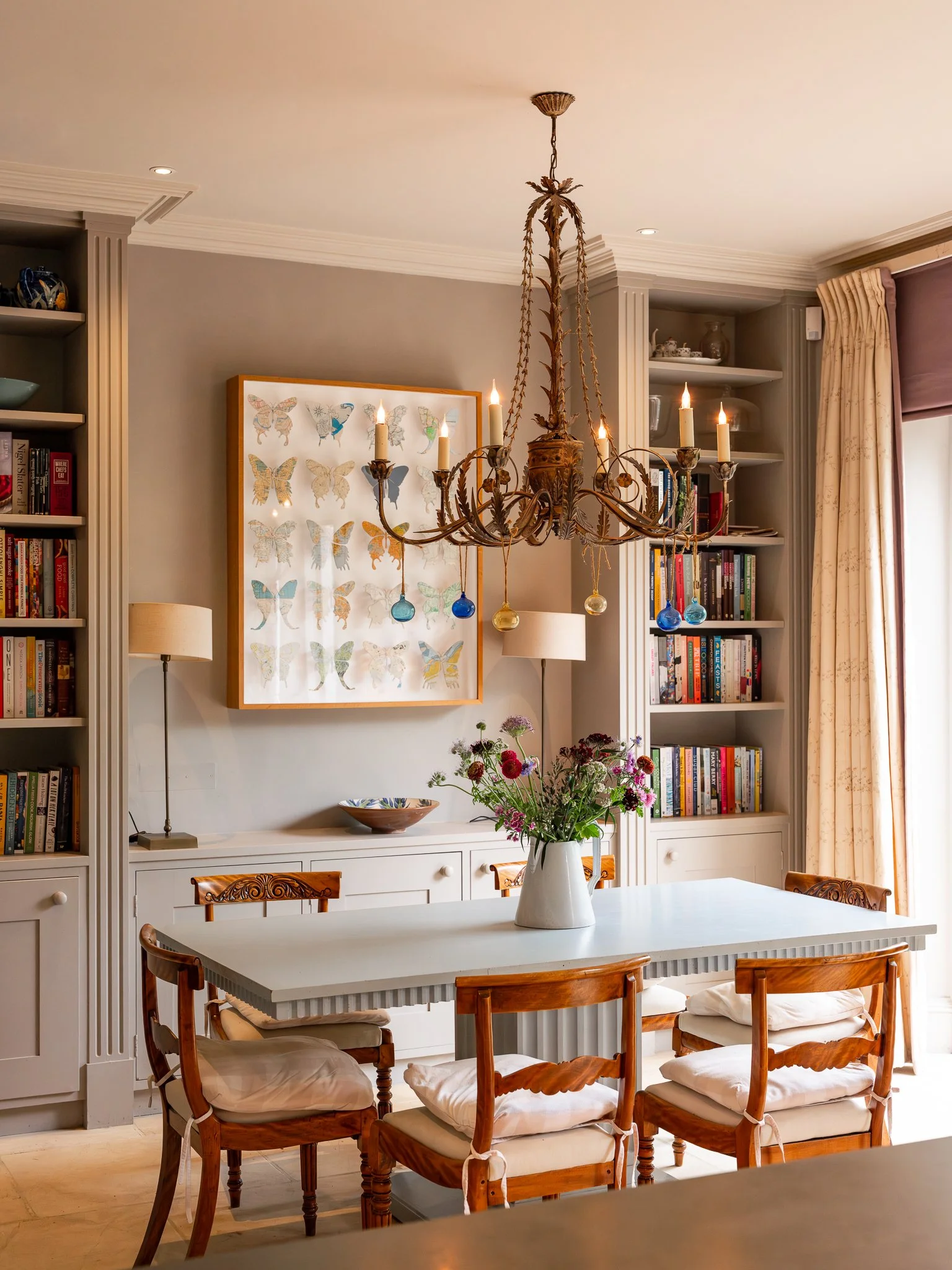 A dining room with a white table, wooden chairs with white cushions, a white sideboard, a vase with flowers, books on shelves, a chandelier with candles, and butterfly artwork on the wall.