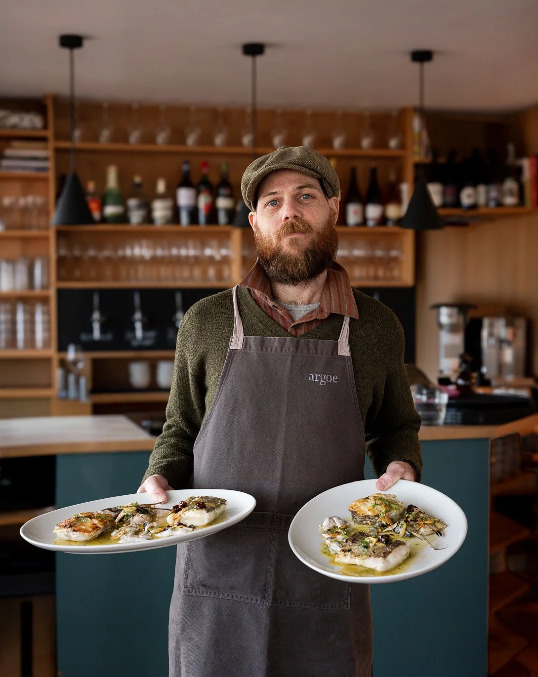 A man with a beard, wearing a newsboy cap, striped shirt, sweater, and apron, holding two plates of food in a kitchen with wooden shelves and bottles in the background.
