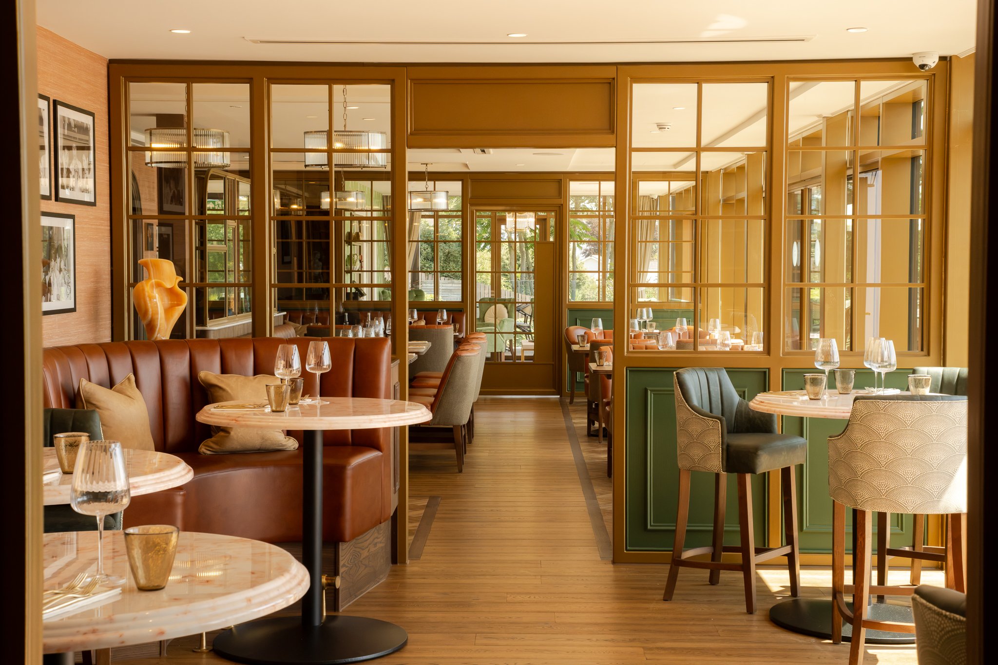 Inside a restaurant with tables and chairs, decorated with framed pictures, a large wooden sculpture, and glassware on the tables. Glass partitions separate different sections of the dining area, with natural light coming through large windows.