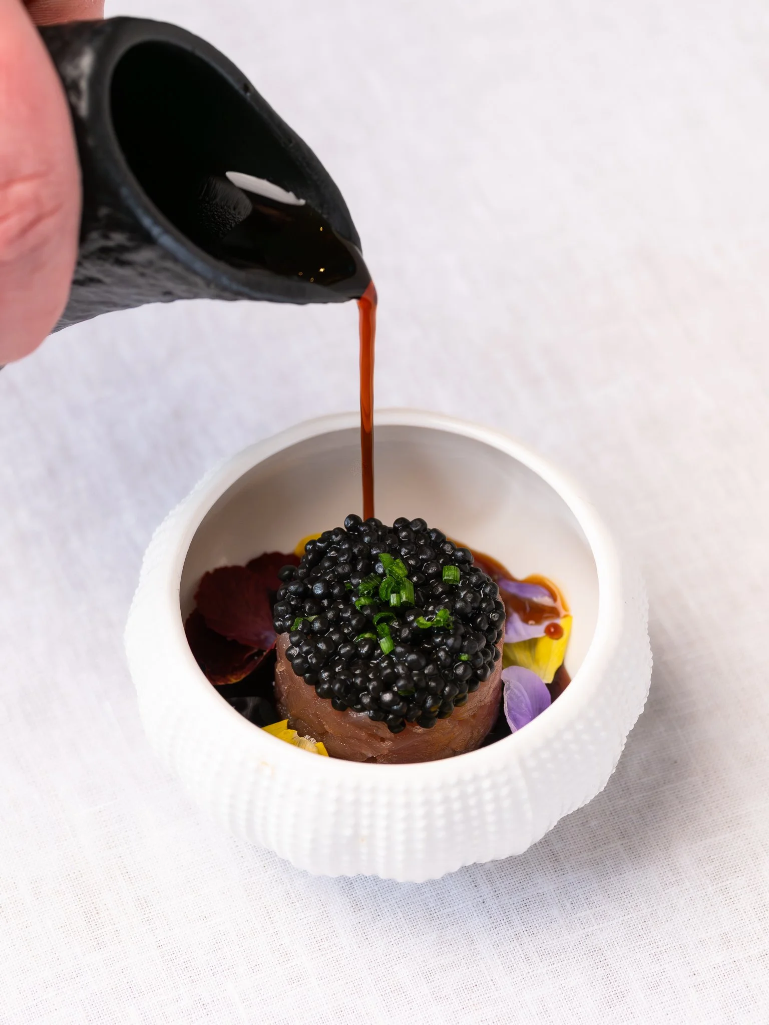 A hand pouring soy sauce over a dish topped with black caviar in a small white bowl, garnished with green onions and edible flower petals on a white tablecloth.