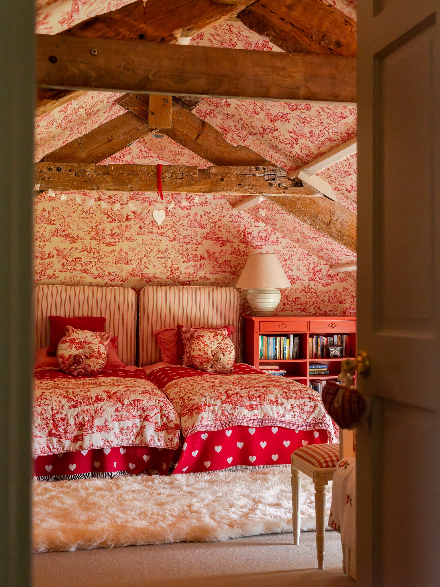 A cozy bedroom with a bed covered in red and white toile patterned bedding, a fluffy cream-colored rug, striped pillows, a red dresser with books and a lamp, and decorated with small heart-themed bedding and a heart-shaped ornament hanging from the c