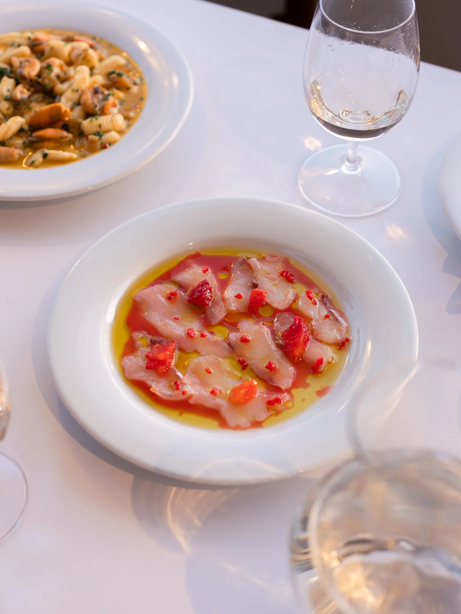 A plate of thinly sliced fish with red pepper and oil, a glass of water, and another plate of pasta with seafood in a creamy sauce on a white table.