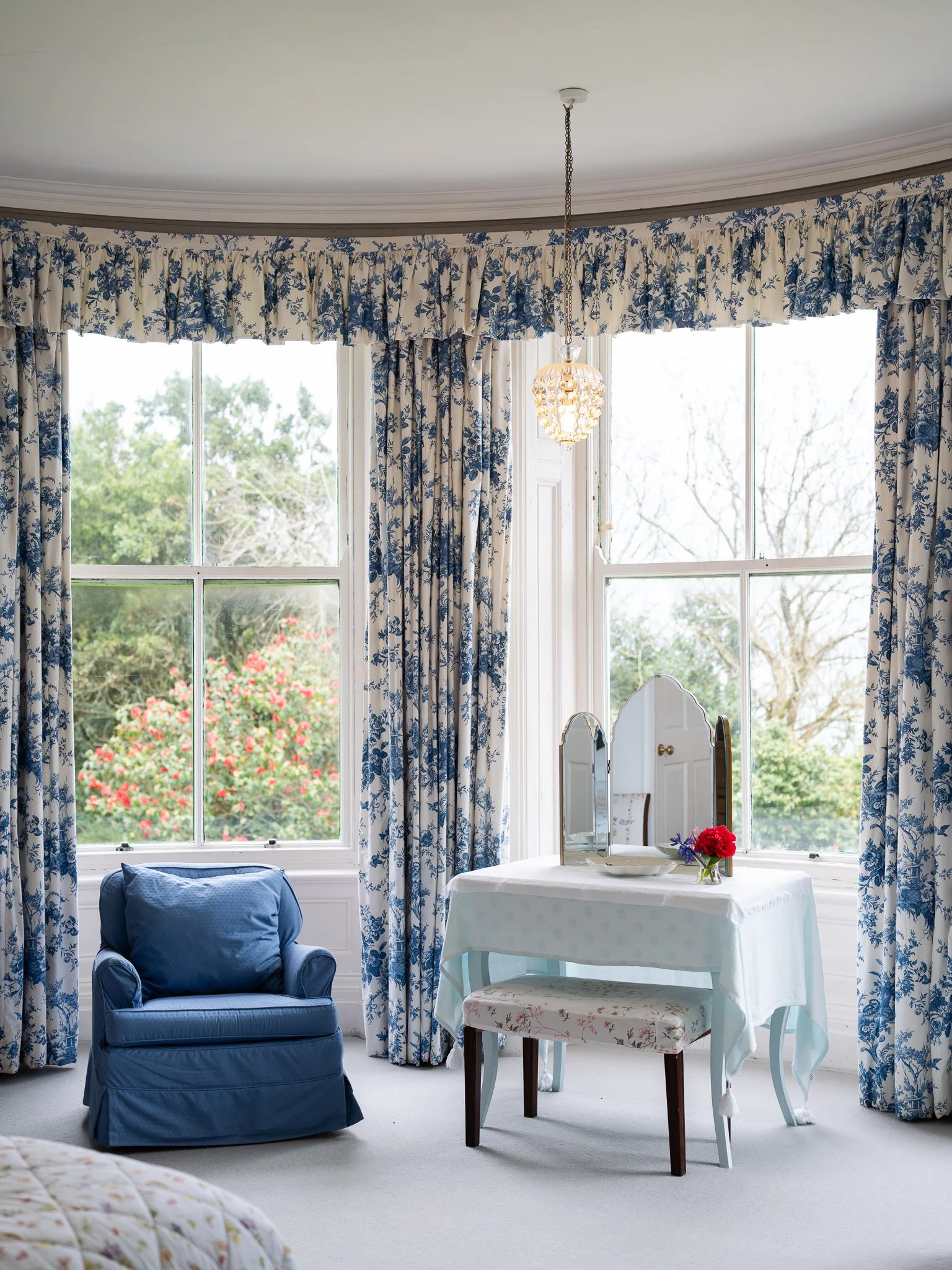 A cozy sunlit room with large windows, blue and white floral curtains, a blue armchair, a vanity with a mirror, a small table with a floral chair, and a vase of red and purple flowers.