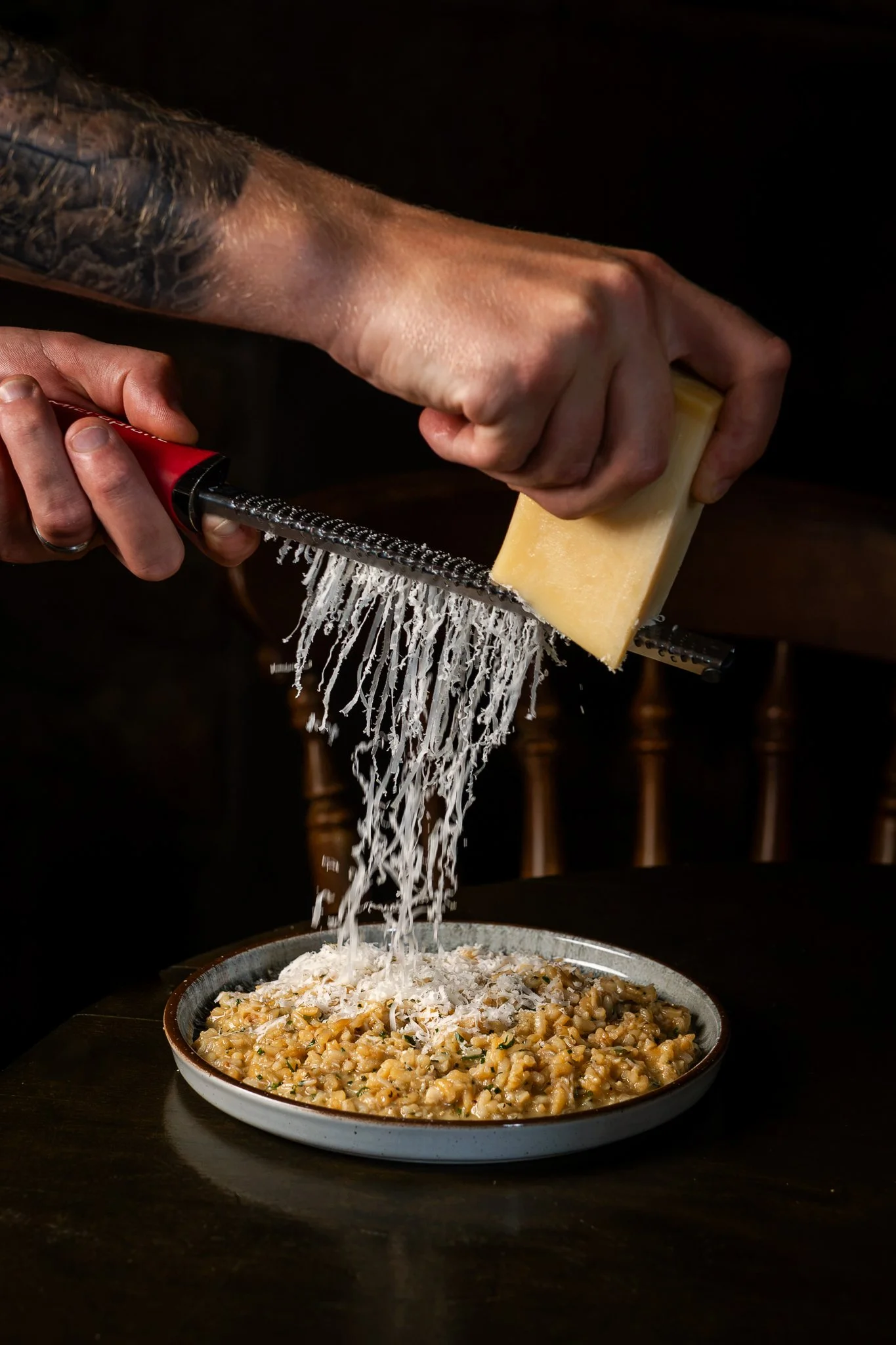 A person grating cheese over a bowl of cooked pasta on a dark table, with a black background.