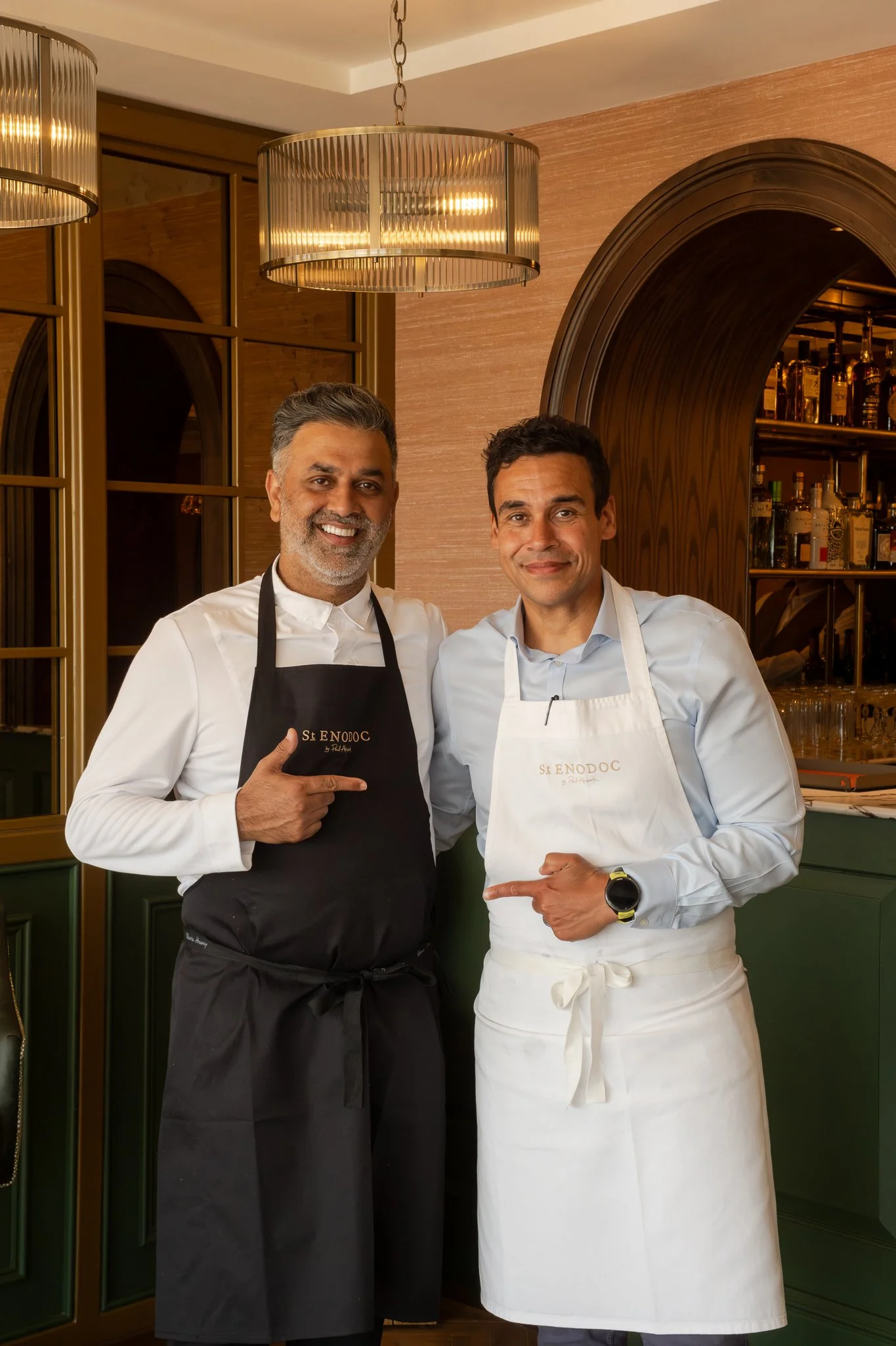 Two male chefs standing together in a restaurant, smiling. One is wearing a black apron and the other a white apron with the words "St ENODOC" on it. They are pointing at each other. The background features a bar with bottles of alcohol and decorativ