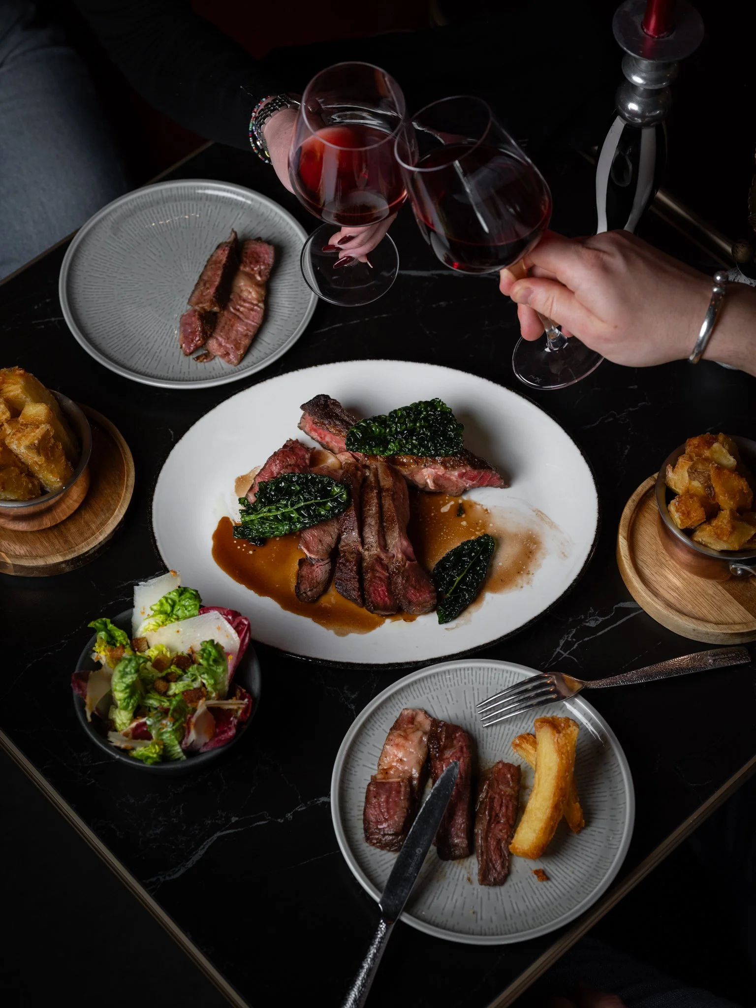 A dinner table with a plate of cooked steak topped with greens and sauce, wine glasses, and various side dishes including salad and potato dishes. Two people are holding glasses of red wine.