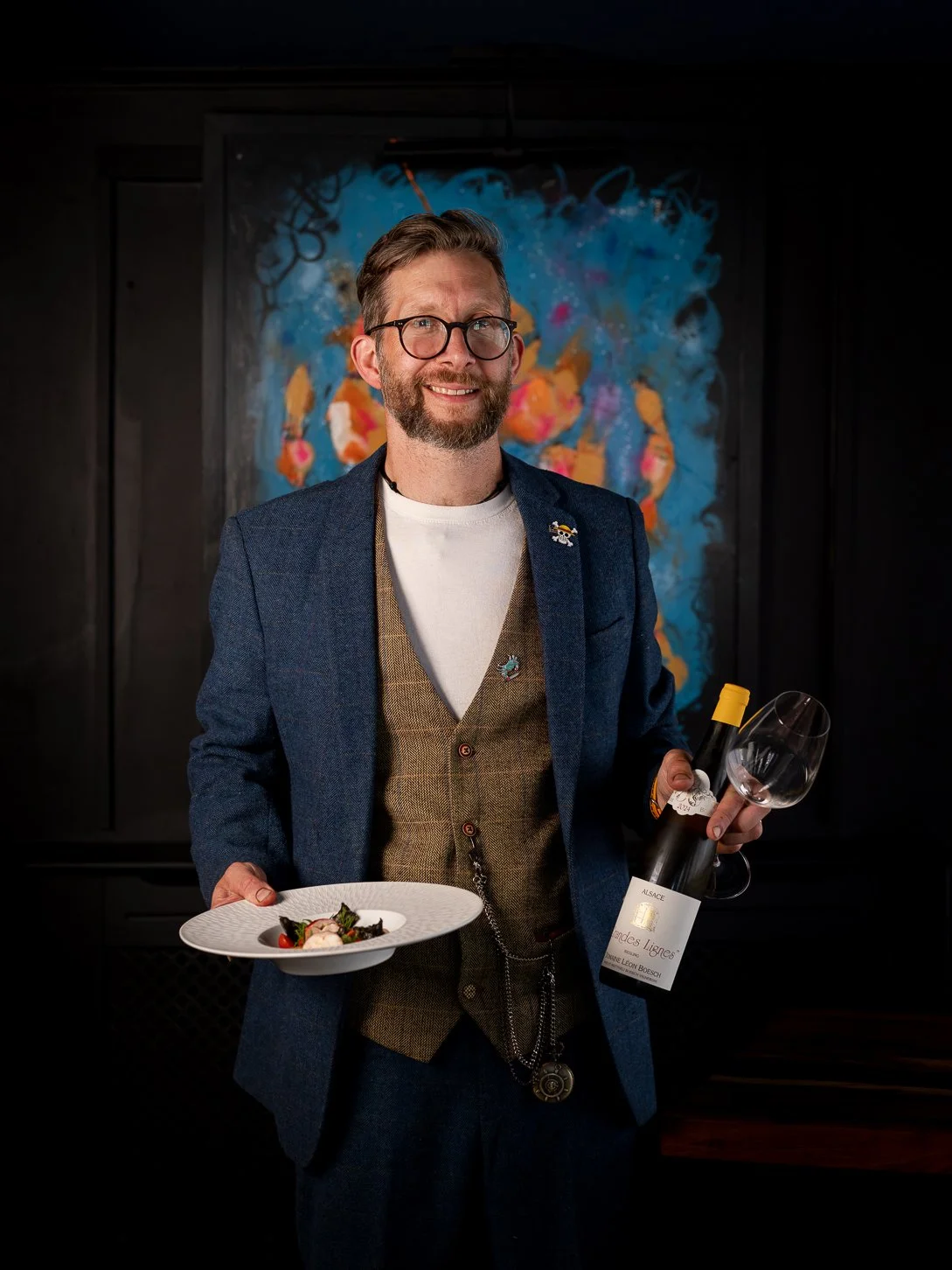 A man with glasses, a beard, and a cheerful expression holding a wine glass and a plate of food, standing in front of an abstract colorful painting.
