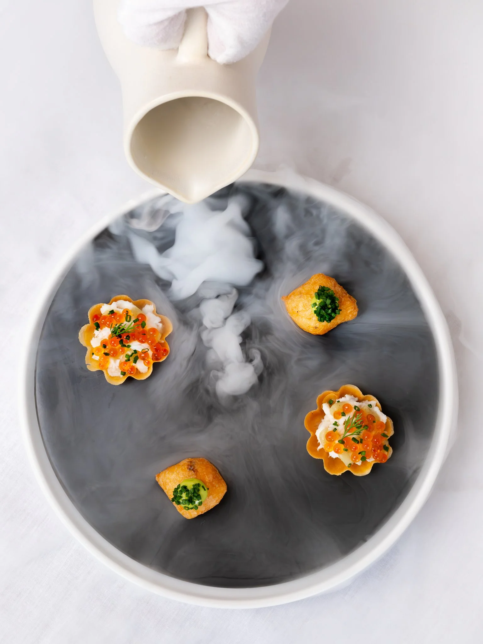 Multiple decorative appetizers topped with orange fish roe, green herbs, and white cream on a circular black plate with swirling smoke or mist. A white pouring vessel is above, releasing smoke or steam.