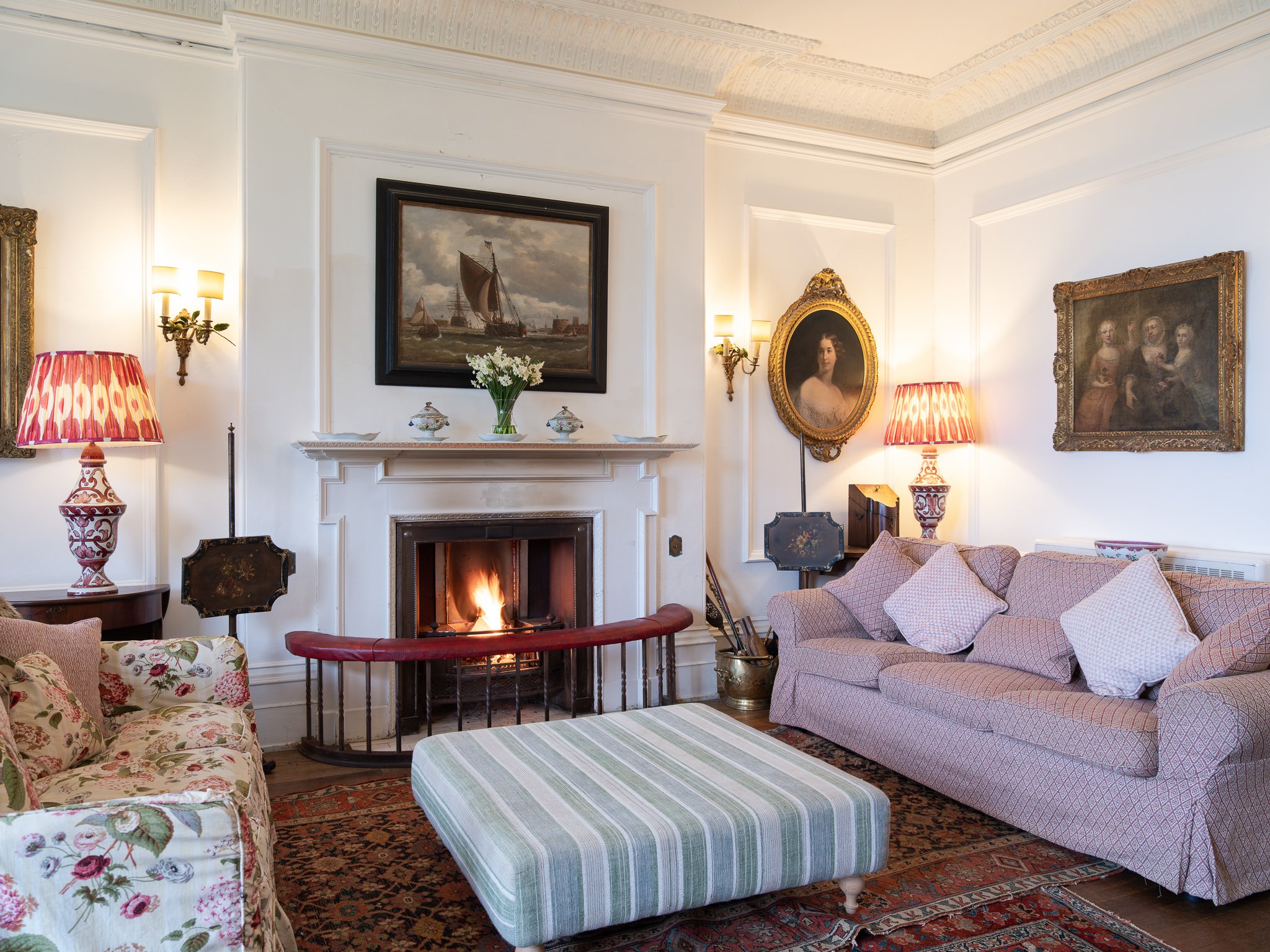 Living room with a lit fireplace, floral sofas, a striped ottoman, framed paintings, and lamps with red and gold shades.