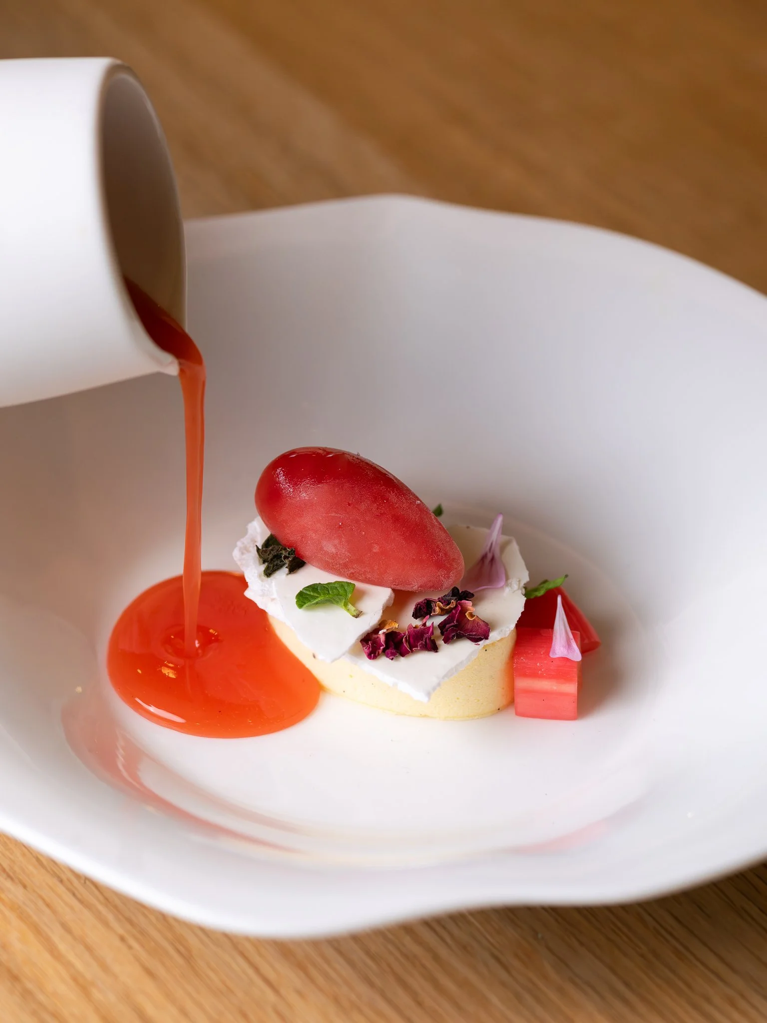 Pouring pink sauce over a dessert with a slice of strawberry, whipped cream, pink flower petals, and edible green garnish on a white plate.