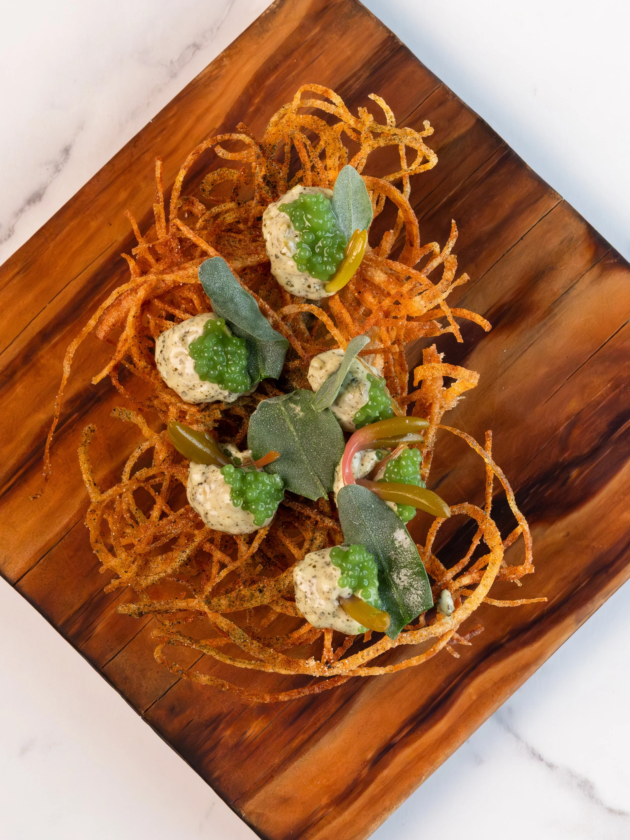 Crispy shredded vegetables topped with dollops of creamy sauce, green caviar, and sage leaves, served on a wooden platter.