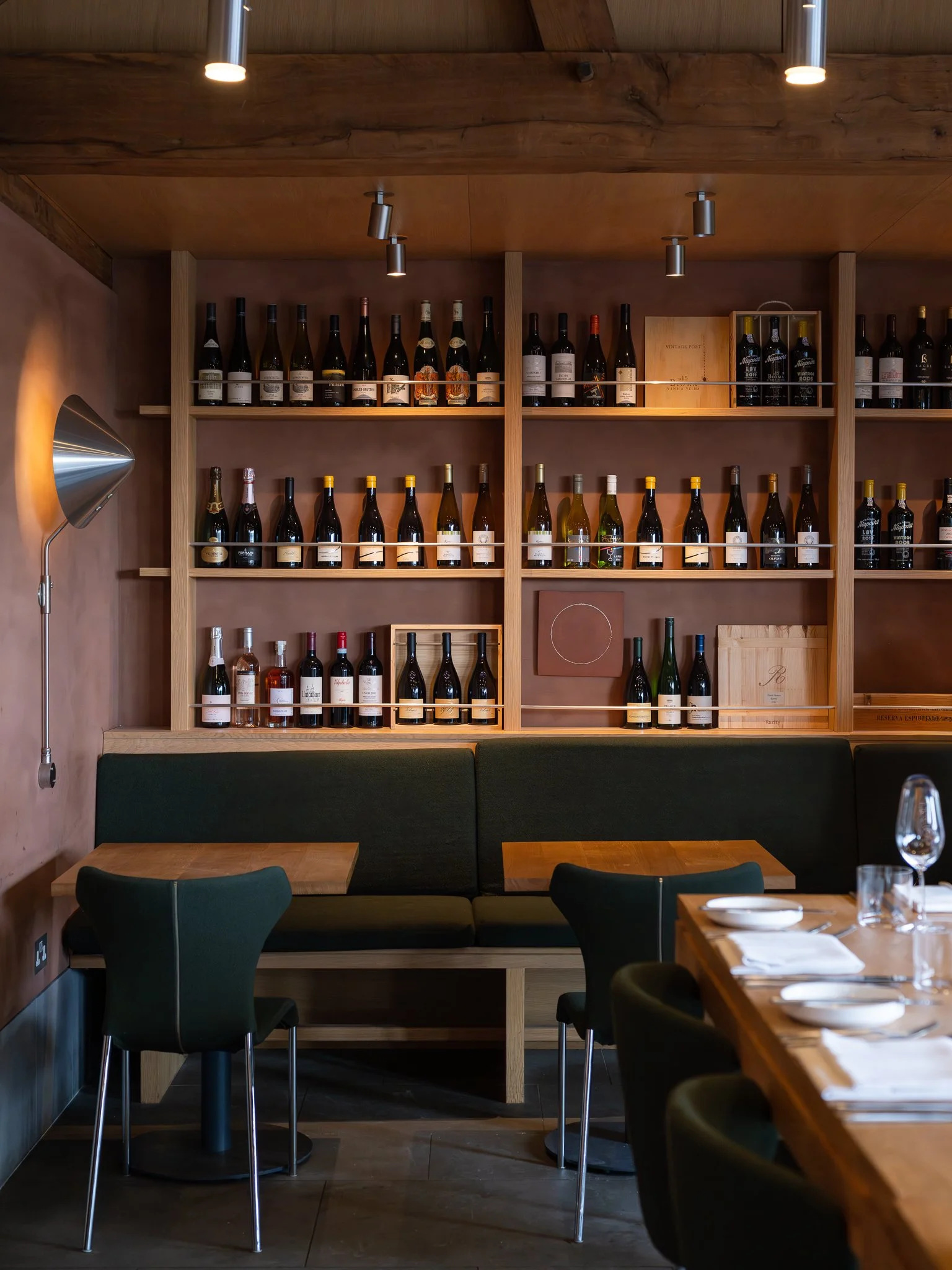 Wine bottles on wooden shelves against a pink wall, with a green upholstered bench and chairs in a restaurant setting