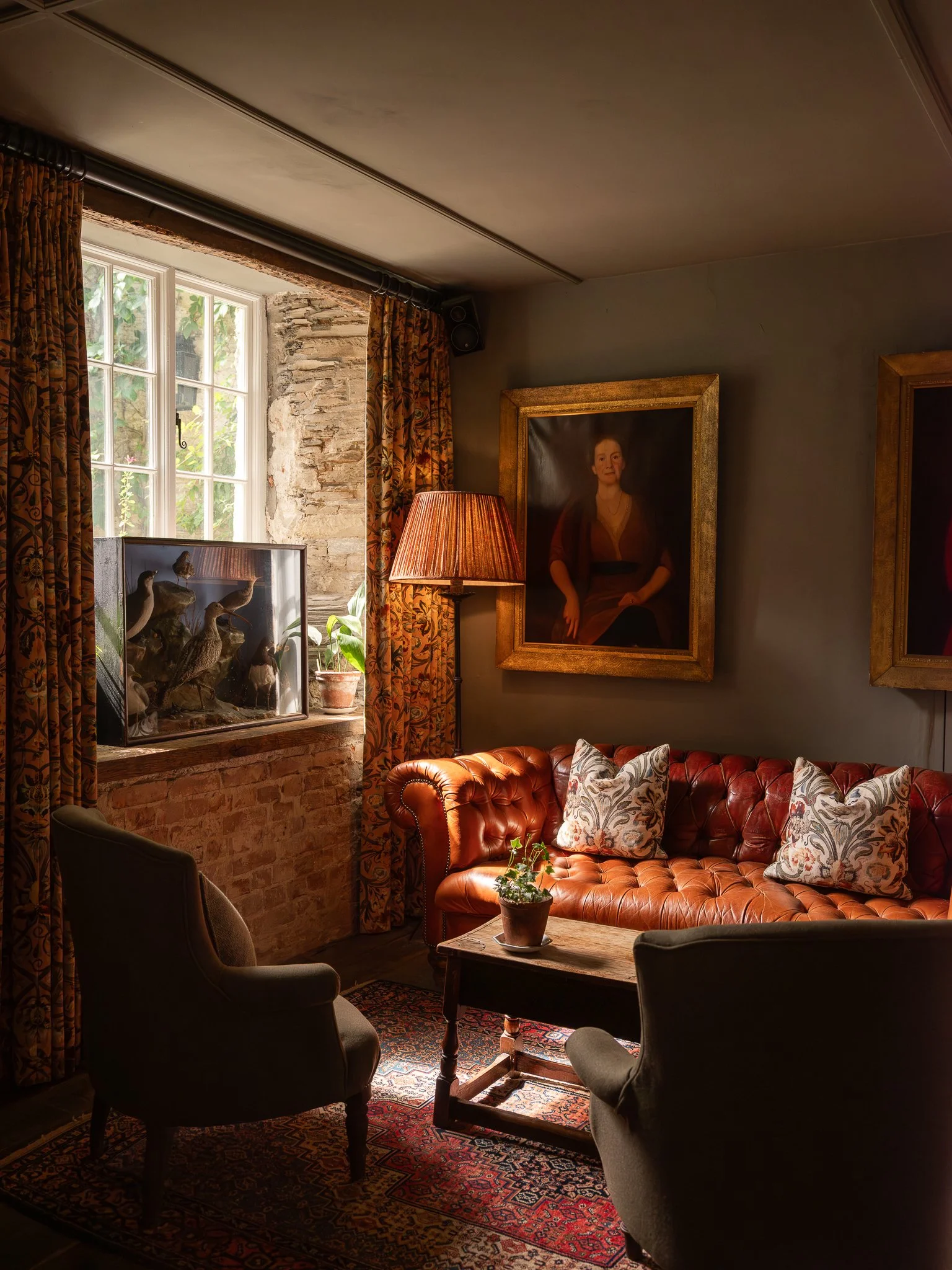 A cozy living room with a leather tufted sofa, patterned cushions, a wooden coffee table with a potted plant, an armchair, a floor lamp with a pleated shade, framed portrait paintings, a window with patterned curtains, a display case with bird figuri