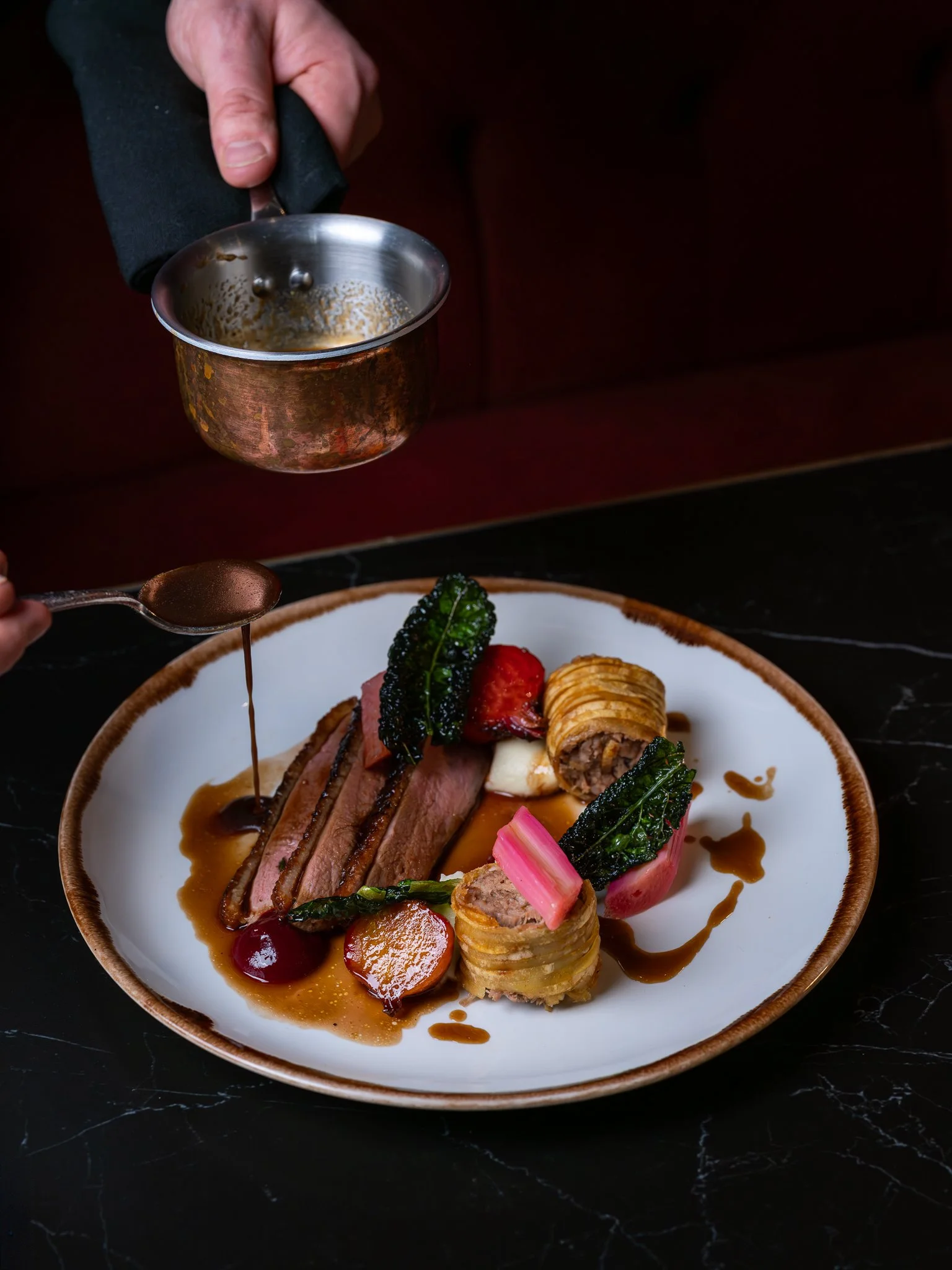 Chef pouring sauce over a gourmet plated dish with sliced steak, vegetables, and sauce