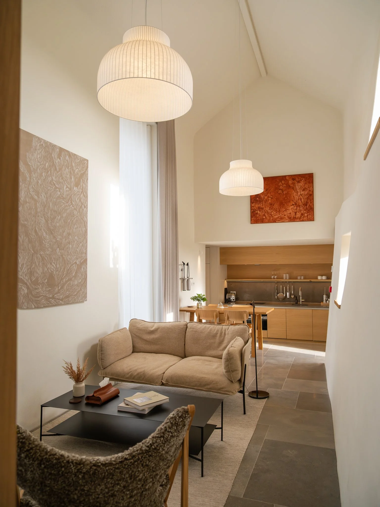 A cozy living room with beige sofa, black coffee table, plush armchair, modern pendant lights, artwork, and a kitchen in the background with a dining table and natural light.