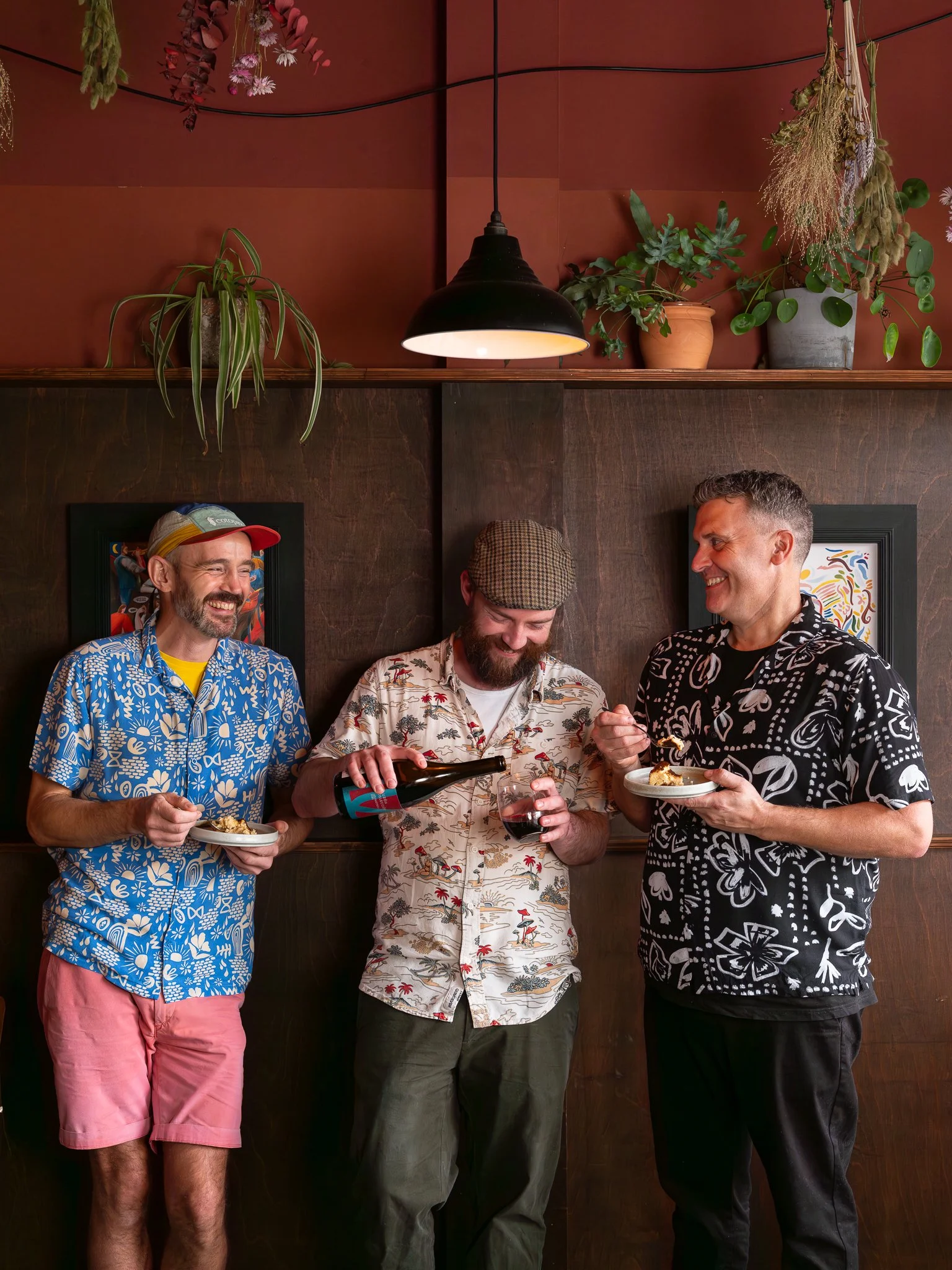 Three men socializing and laughing in a restaurant. They are wearing patterned shirts, and one is holding a plate of food, another holding a glass of wine, and the third pouring a drink.