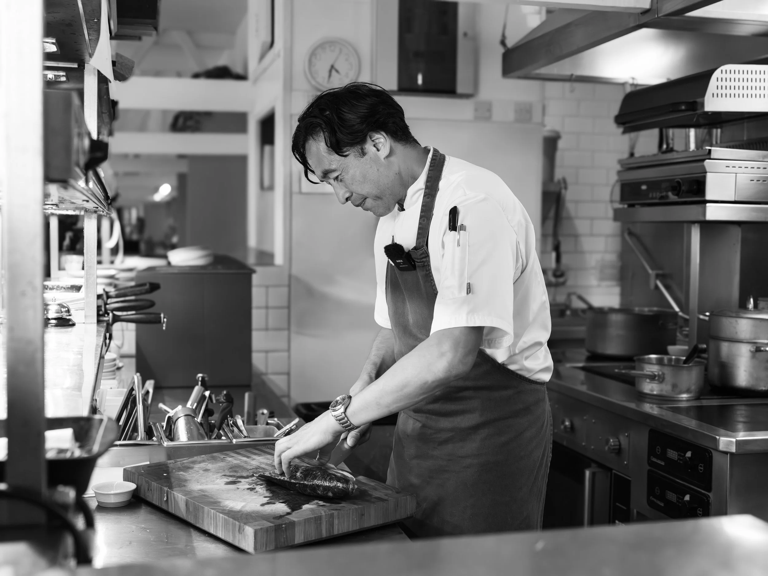 A chef in a commercial kitchen preparing a fish on a wooden cutting board, surrounded by kitchen equipment and utensils.