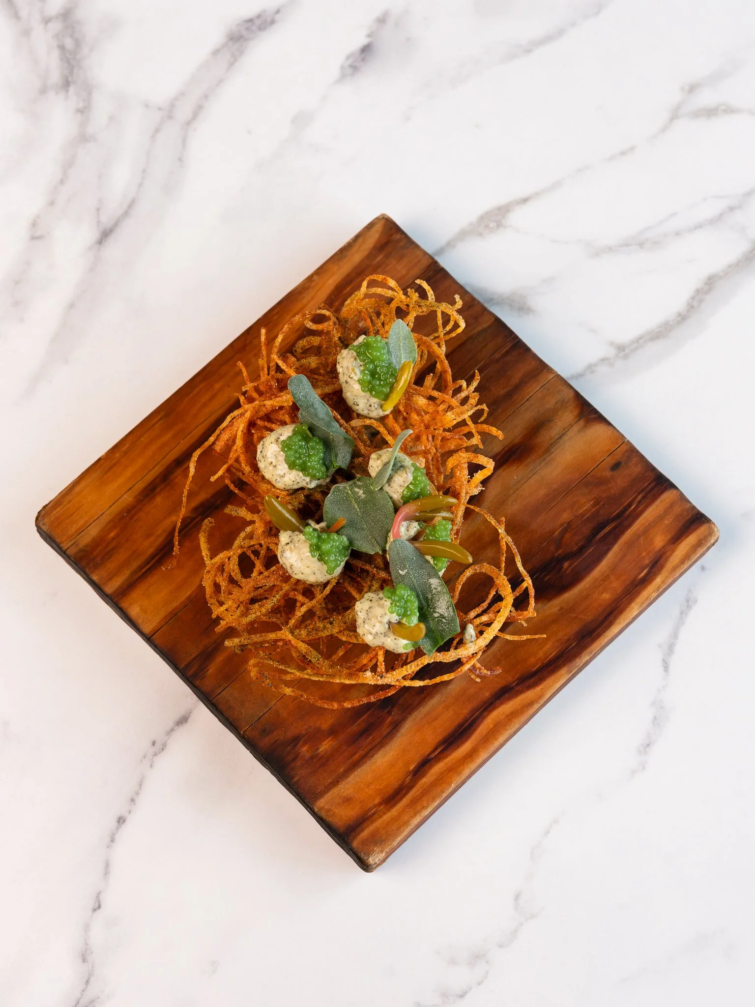 A dish featuring crispy fried onion rings garnished with green sauce, herbs, and small greenery, served on a wooden plate.