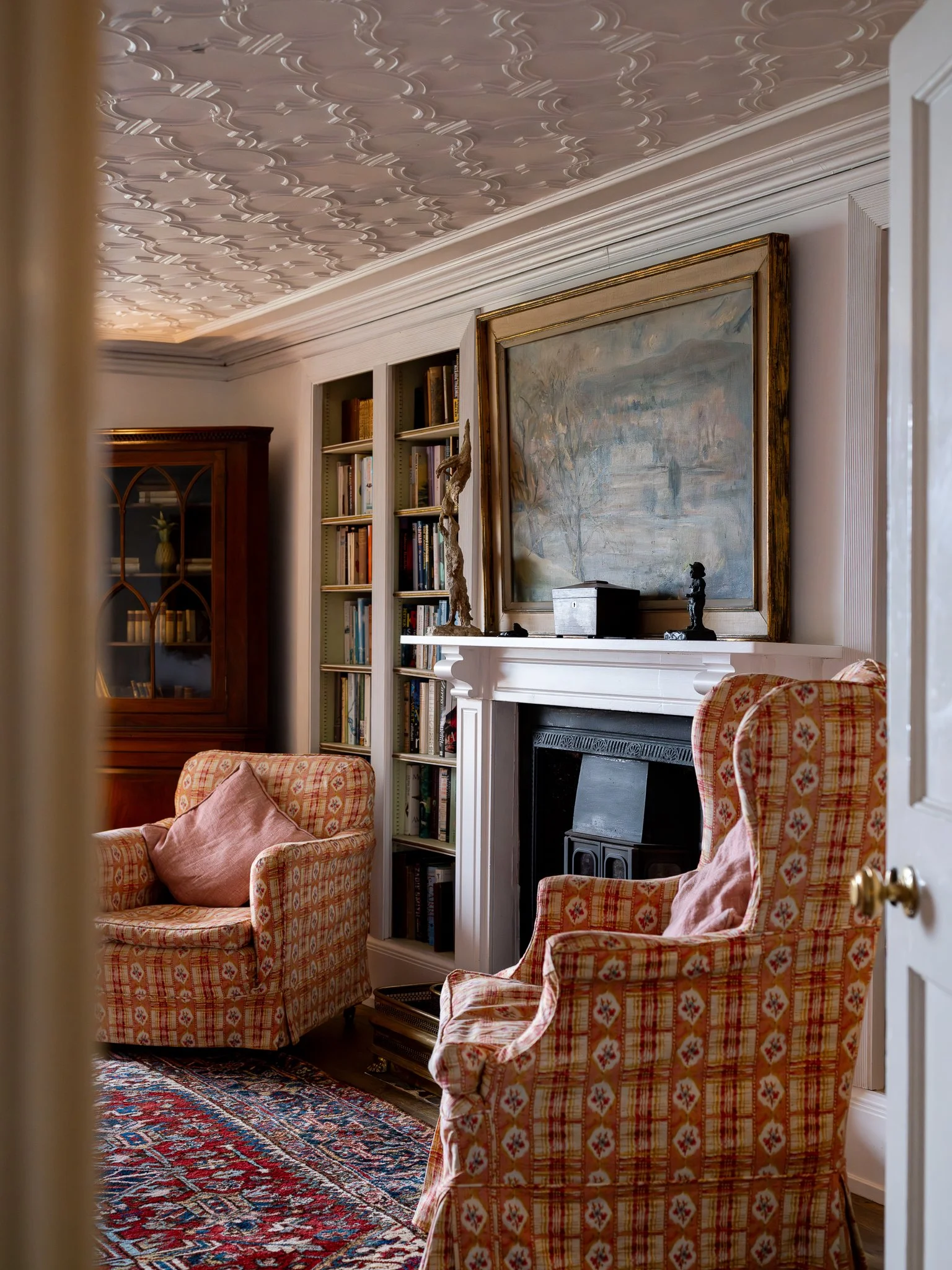 Cozy living room with patterned armchairs, a white fireplace, framed landscape painting, bookshelf, and decorative ceiling molding.