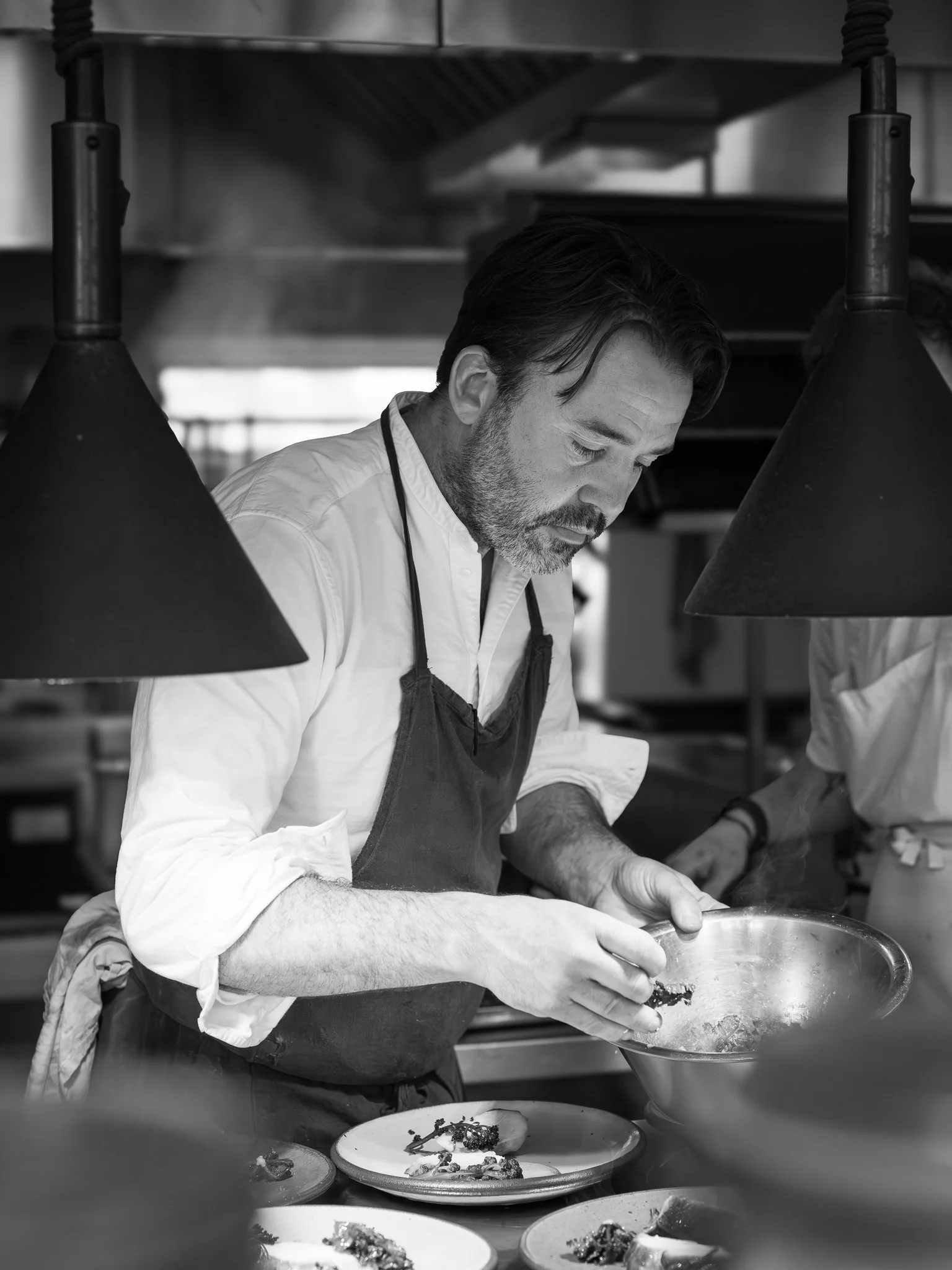 A chef with a beard and mustache wearing a white shirt and an apron, focused on plating dishes in a professional kitchen. Several plates with food can be seen on the counter in front of him.