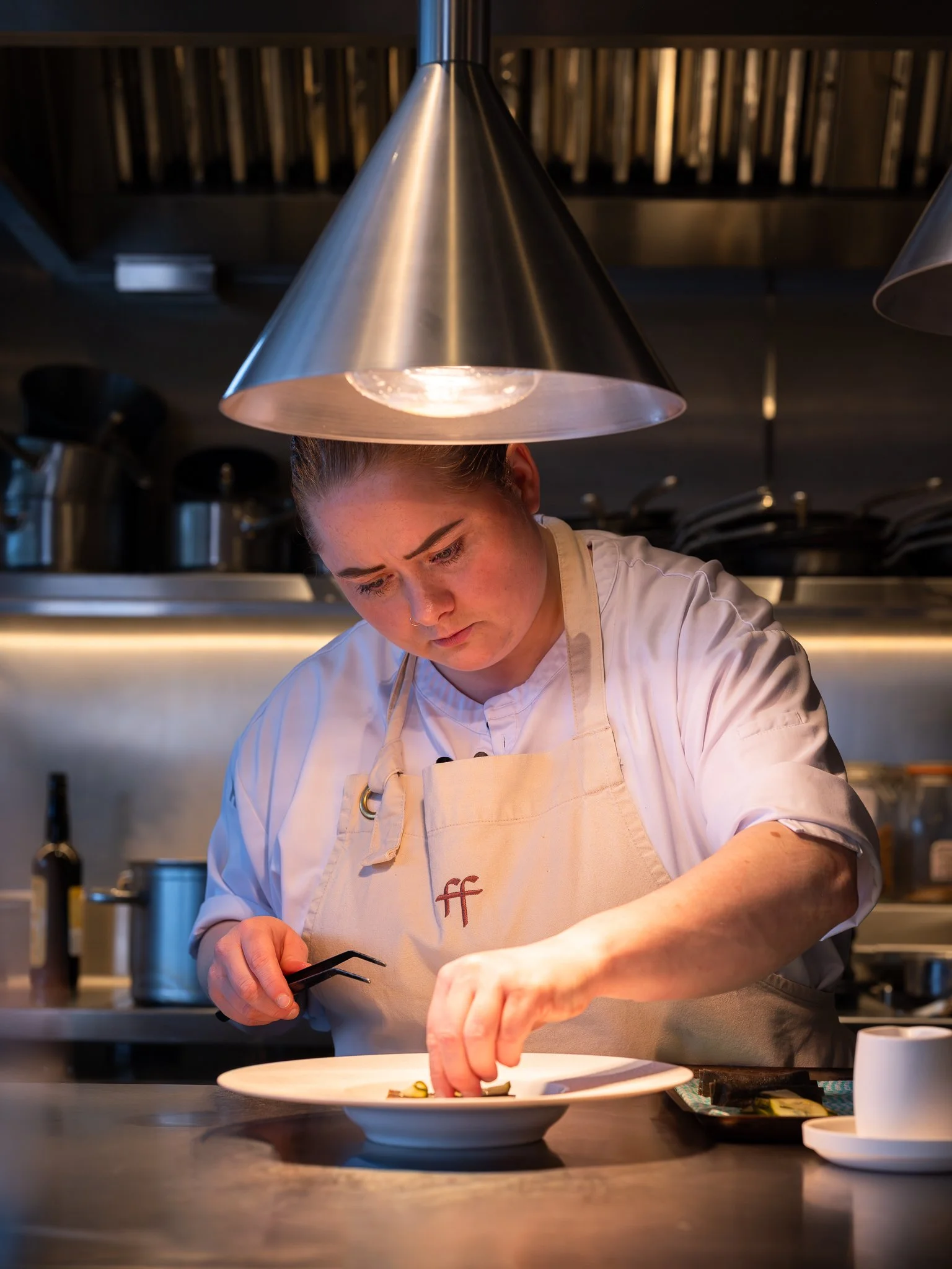 A chef carefully plates food in a professional kitchen, wearing a white coat and beige apron under large metallic restaurant lamps.