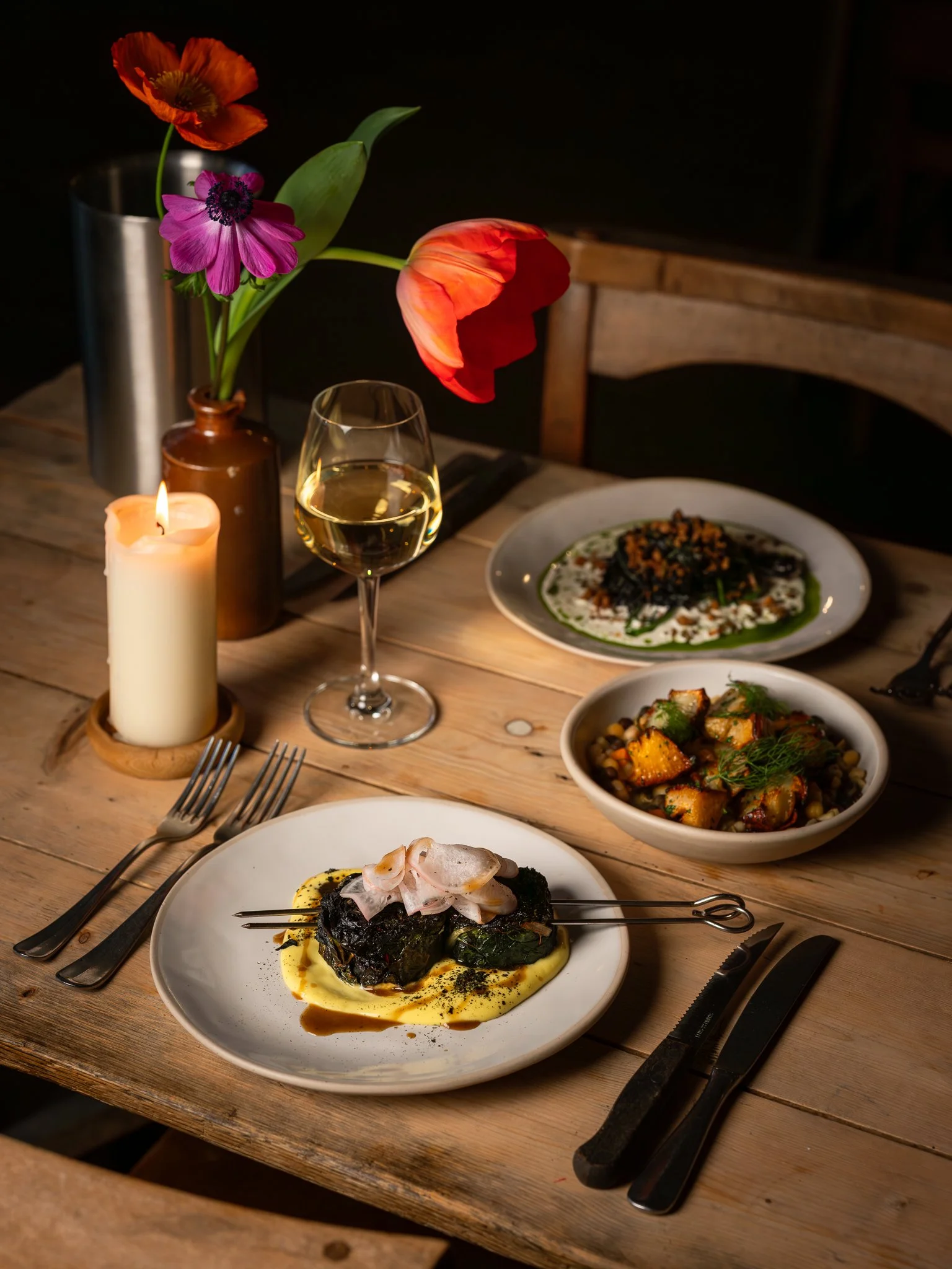 A dinner table with a flower arrangement, a candle, a glass of white wine, and various dishes including a plated entree and bowls of vegetables and grains.