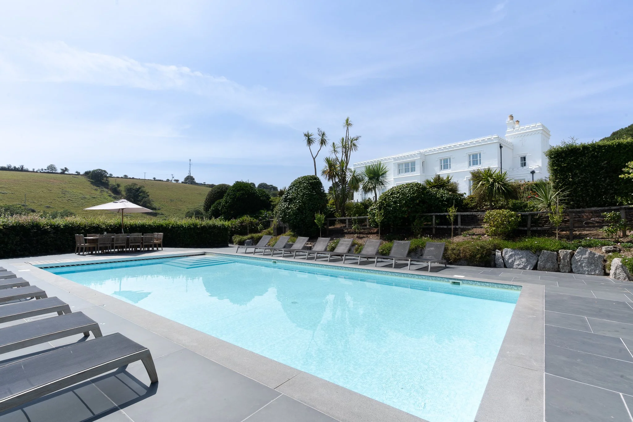 Outdoor swimming pool with lounge chairs, a white house, scenic hills, and a clear blue sky.