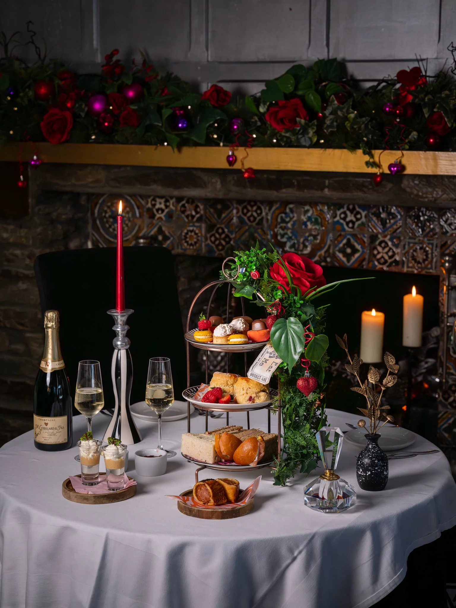 Elegant holiday dessert table with champagne, macarons, cakes, and chocolates, decorated with a red rose and greenery, set on a round table with white tablecloth, candles, and festive garland in the background.