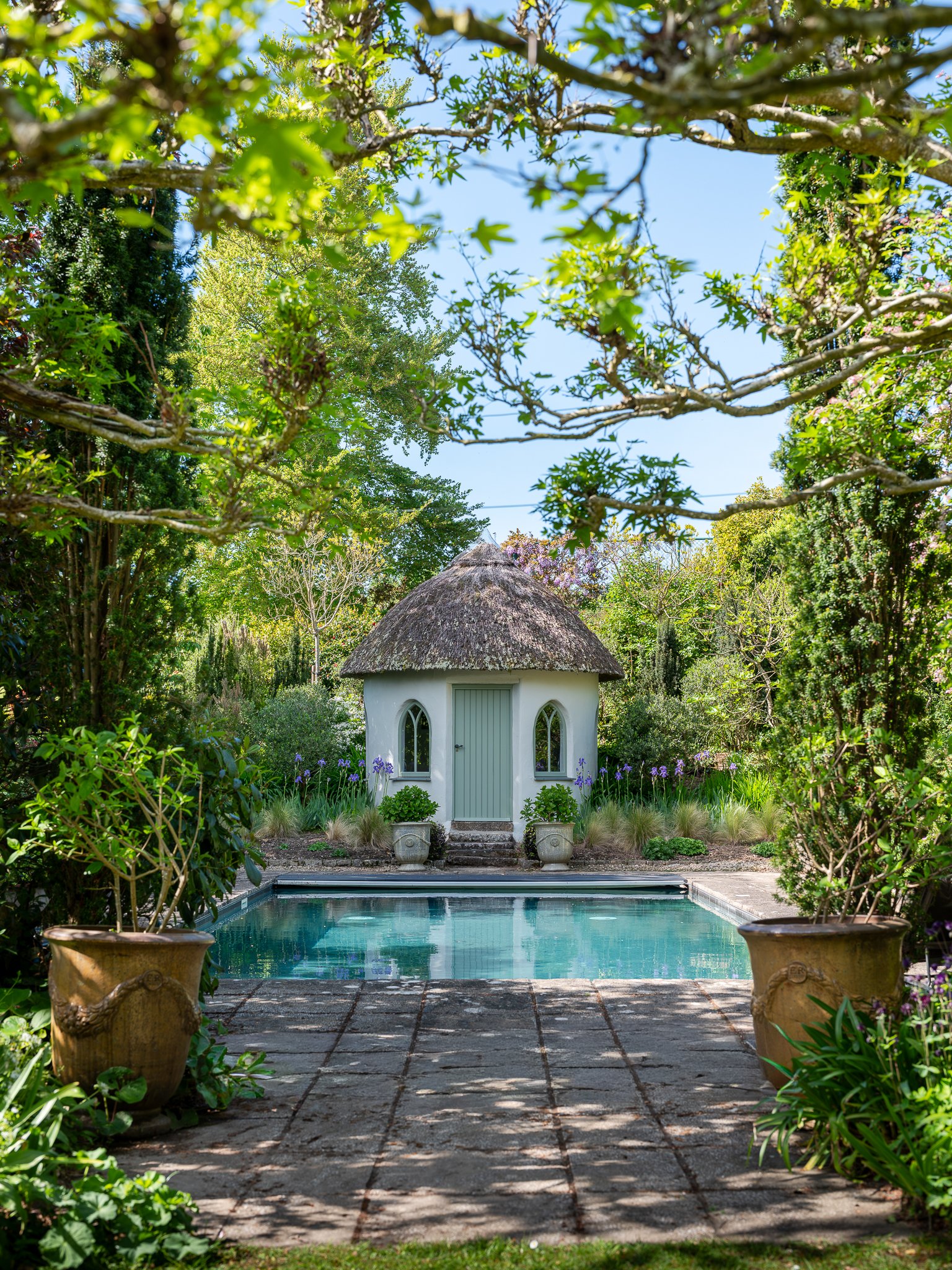 A small white house with a thatched roof, two arched windows, and a light blue door, set in a lush garden with green foliage, potted plants, and a swimming pool in the foreground, under a partly cloudy sky.
