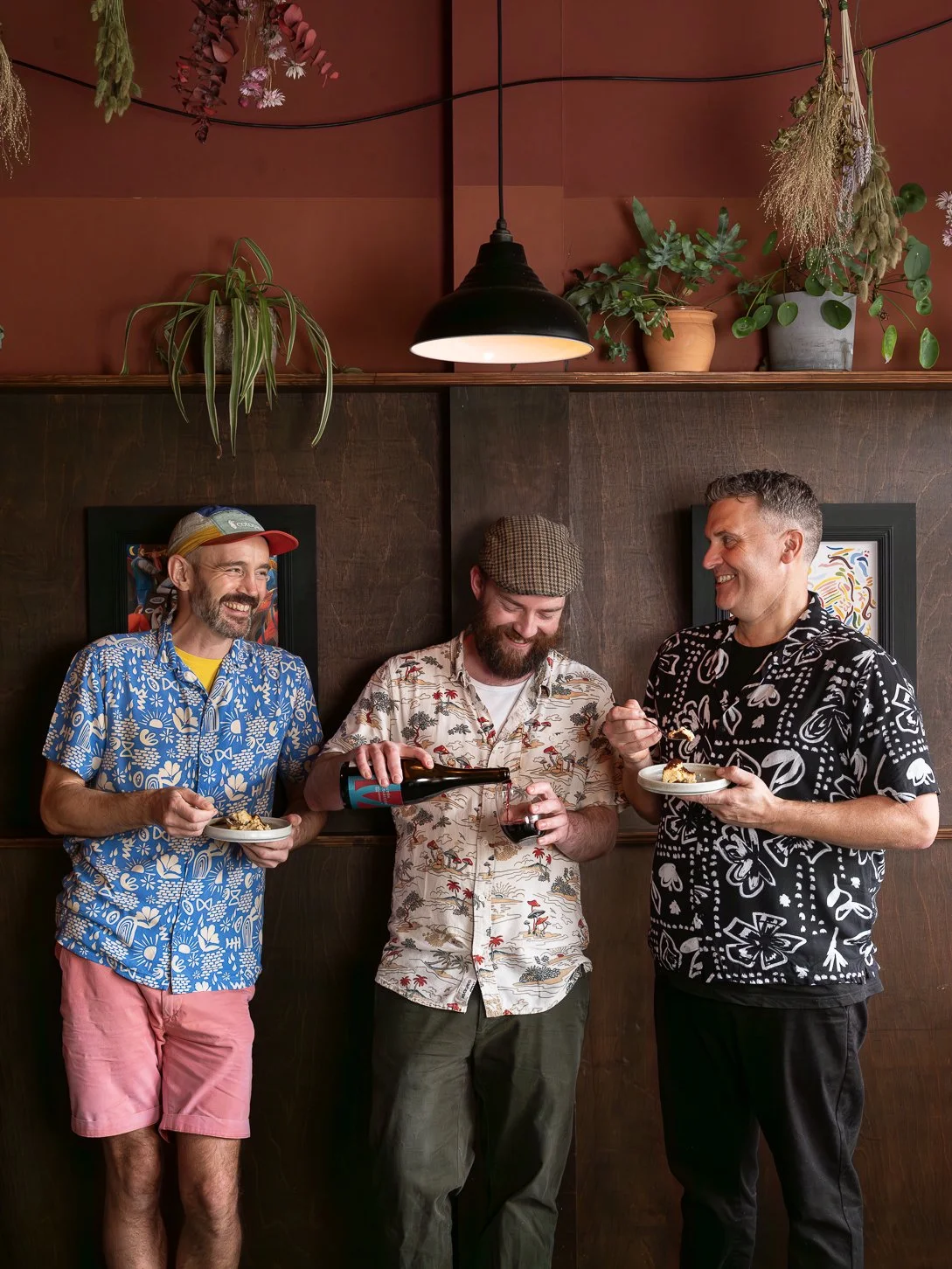 Three men in colorful Hawaiian shirts enjoying food and wine in an indoor setting with plants and artwork on the wall.