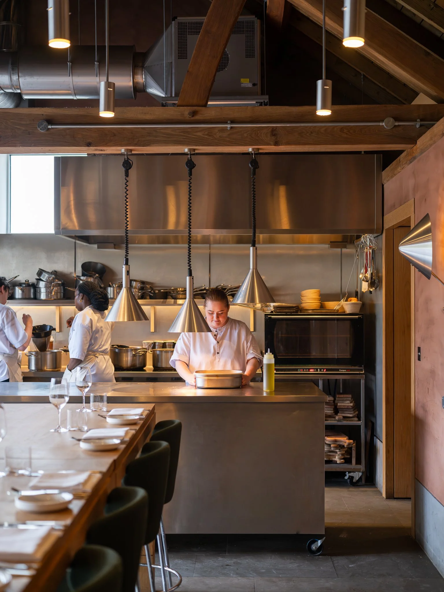 Kitchen scene with three chefs working behind a counter in a restaurant, stainless steel appliances, and wooden accents.