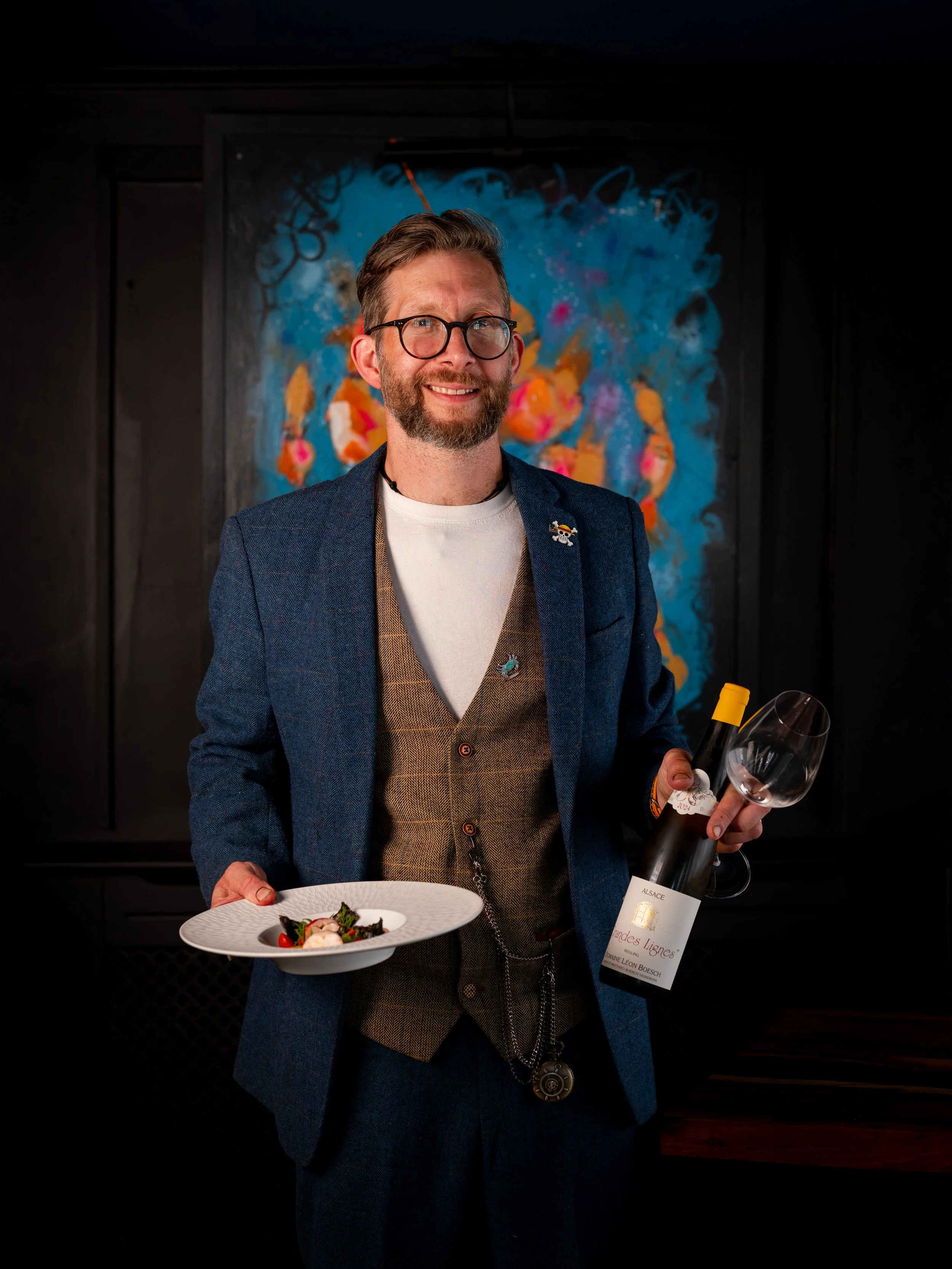 A man with glasses and a beard is smiling while holding a plate of food and a bottle of wine in a dimly lit room with a colorful abstract painting in the background.