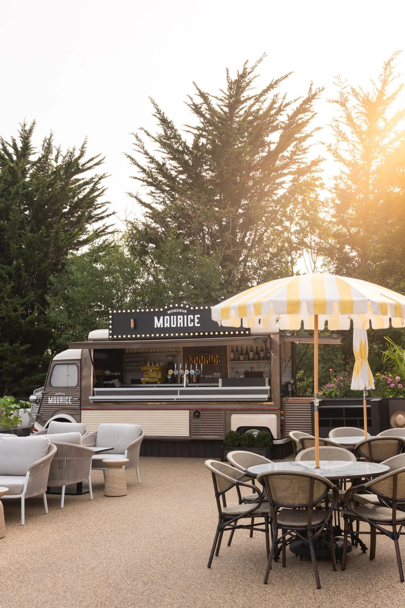 Outdoor setting with a vintage food truck named Monsieur Maurice serving drinks, surrounded by white and black wicker chairs, a yellow and white striped umbrella, and lush green trees in the background, with warm sunlight filtering through.