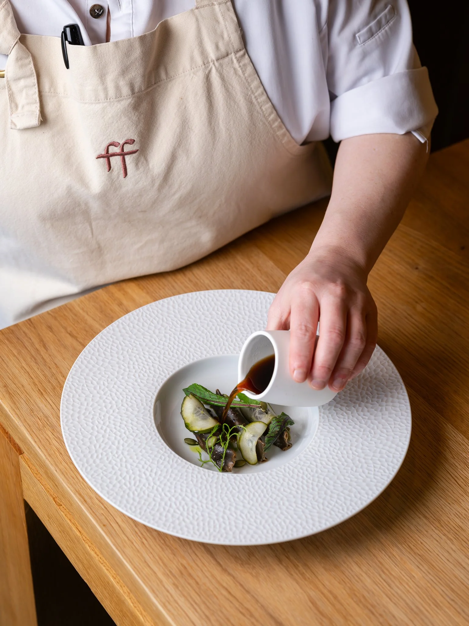A chef pouring soy sauce over a dish of vegetables and garnishes on a textured white plate.