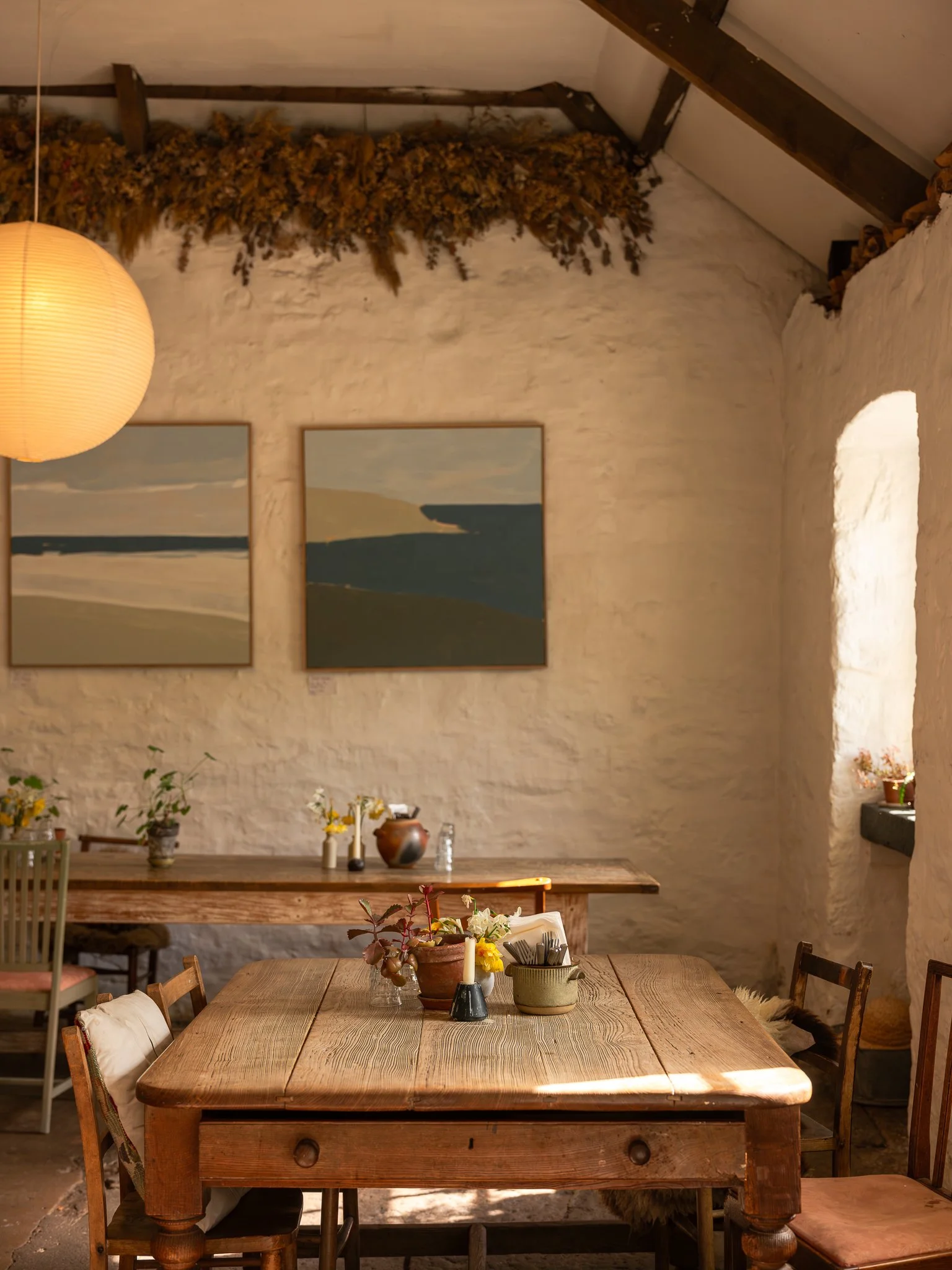 A cozy dining area featuring a large wooden table with decorative items including candles and flowers in pots. The room has a rustic feel with stone walls, framed landscape artwork, and warm lighting from a paper lantern. There are chairs around the 
