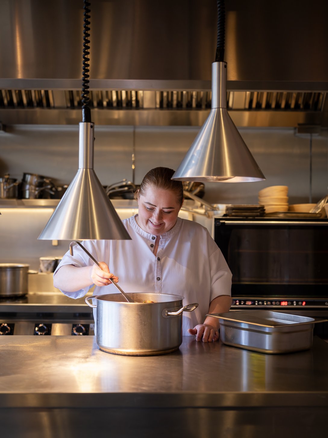 A female chef smiling and stirring a large silver pot in a professional kitchen, with stainless steel appliances and utensils in the background.