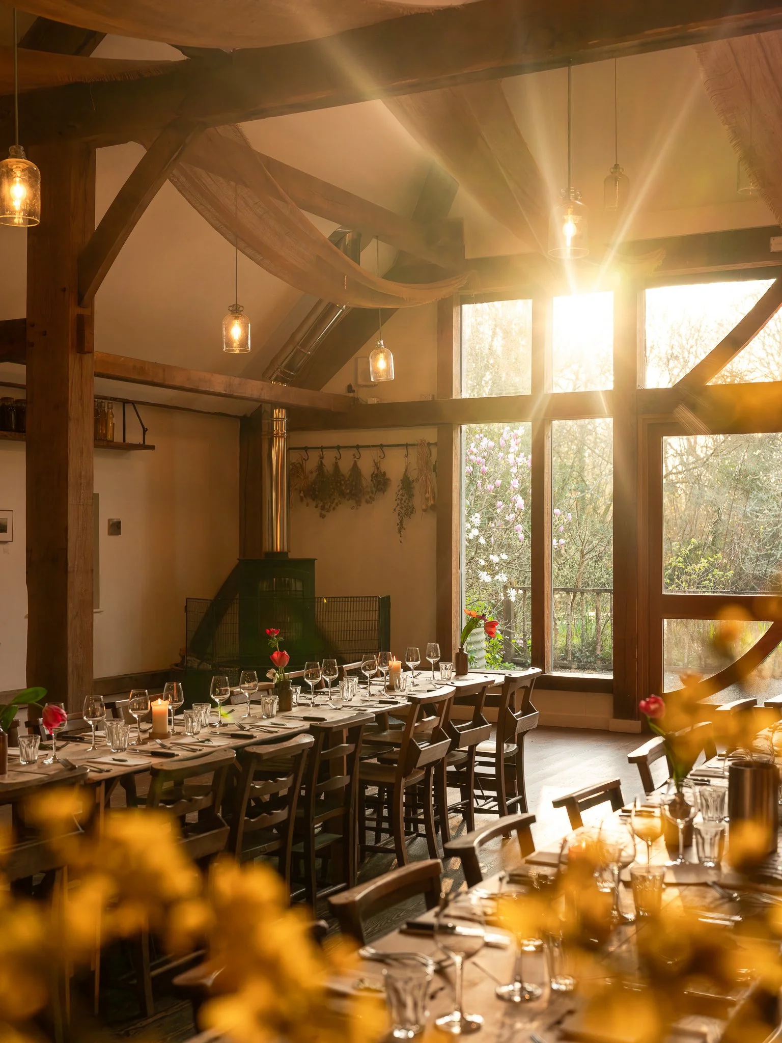 A dining room with a long wooden table set with glasses and candles, illuminated by warm sunlight through large windows and decorated with flowers.