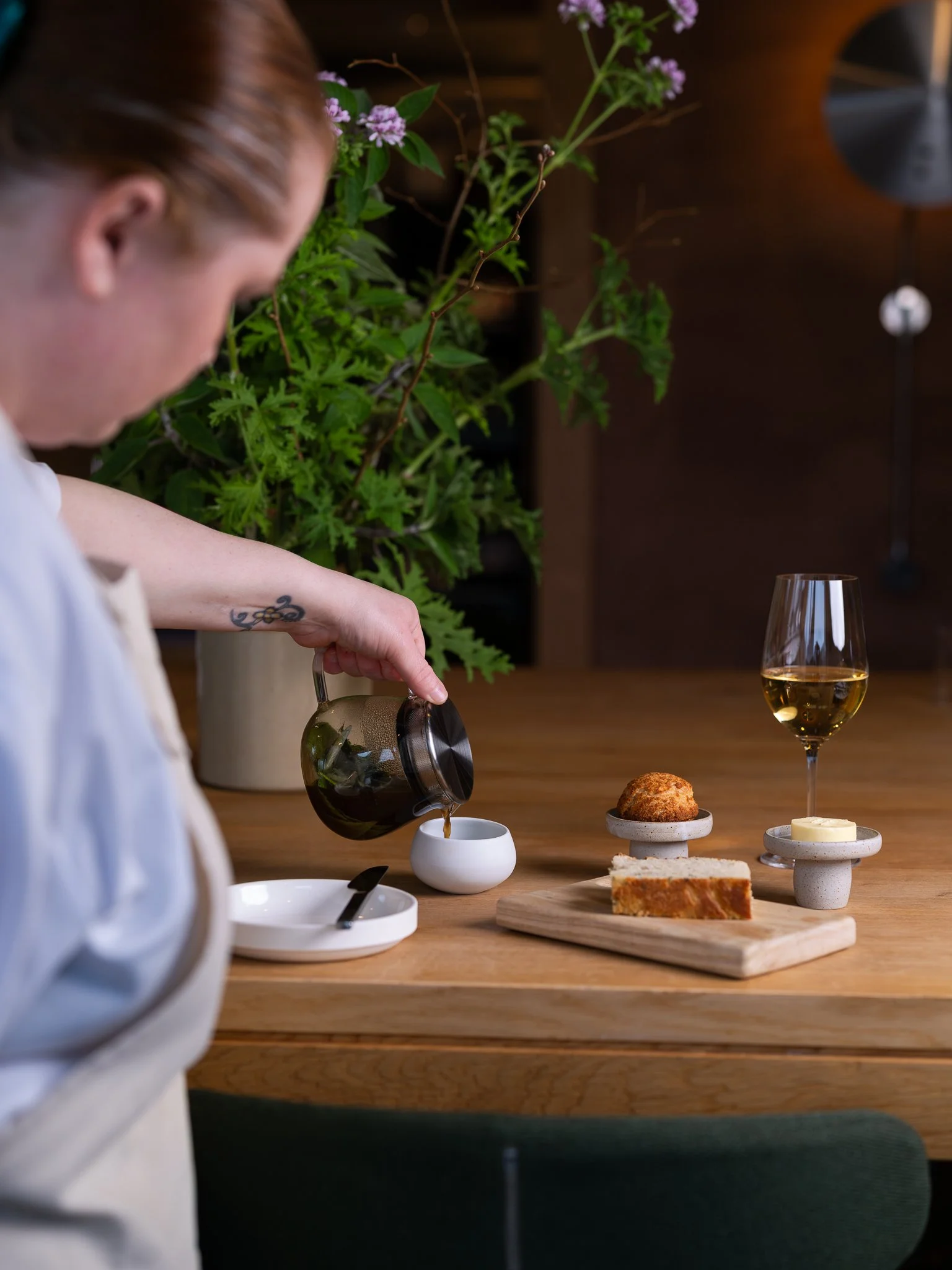 Person pouring coffee or tea at a wooden table with a glass of white wine, a small cake, a slice of cake, and small serving dishes, and leafy green plants in the background.